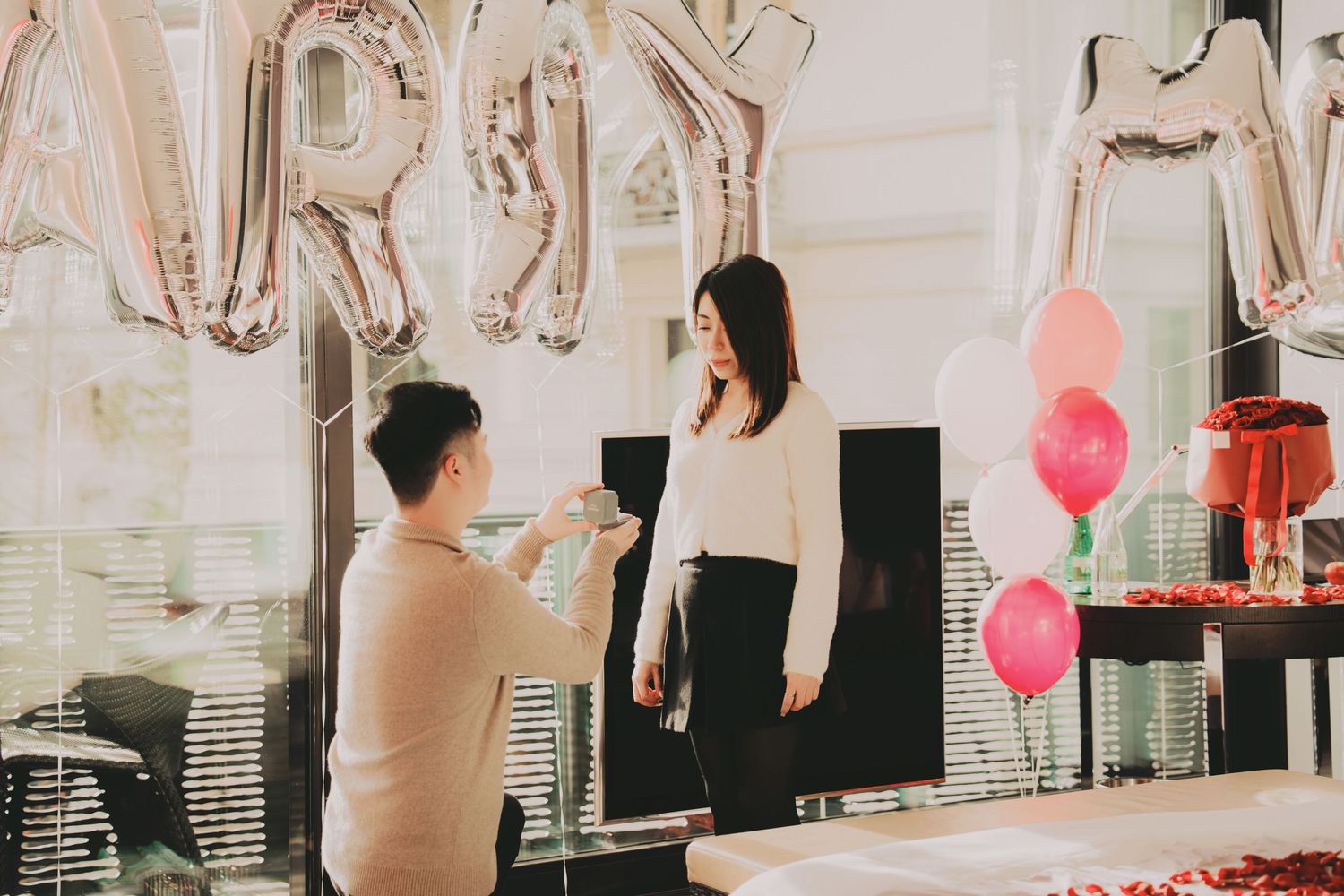 Man proposing to his girlfriend in a festive room decorated with silver MARRY balloons and pink heart balloons.