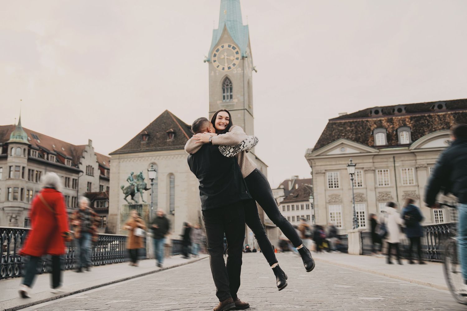 Man lifting woman joyfully on a busy Zurich bridge with historic architecture behind.