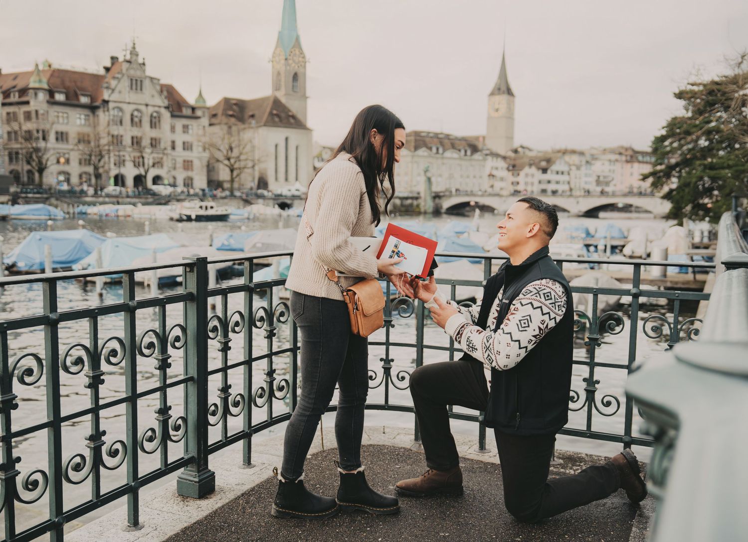 Romantic marriage proposal on Zurich bridge with man kneeling holding ring box and gift.