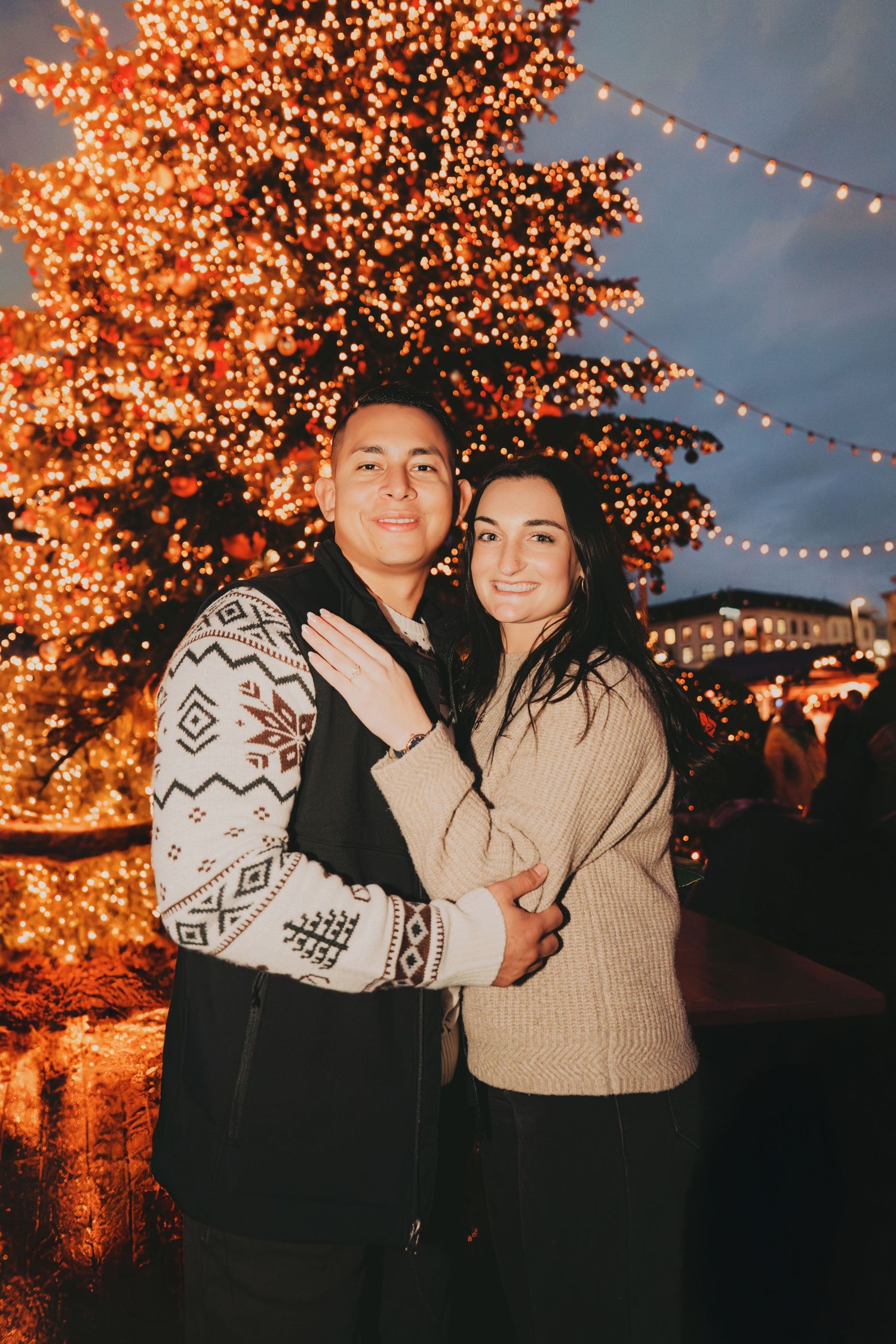 Happy couple embracing in front of a beautifully lit Christmas tree at a holiday market.