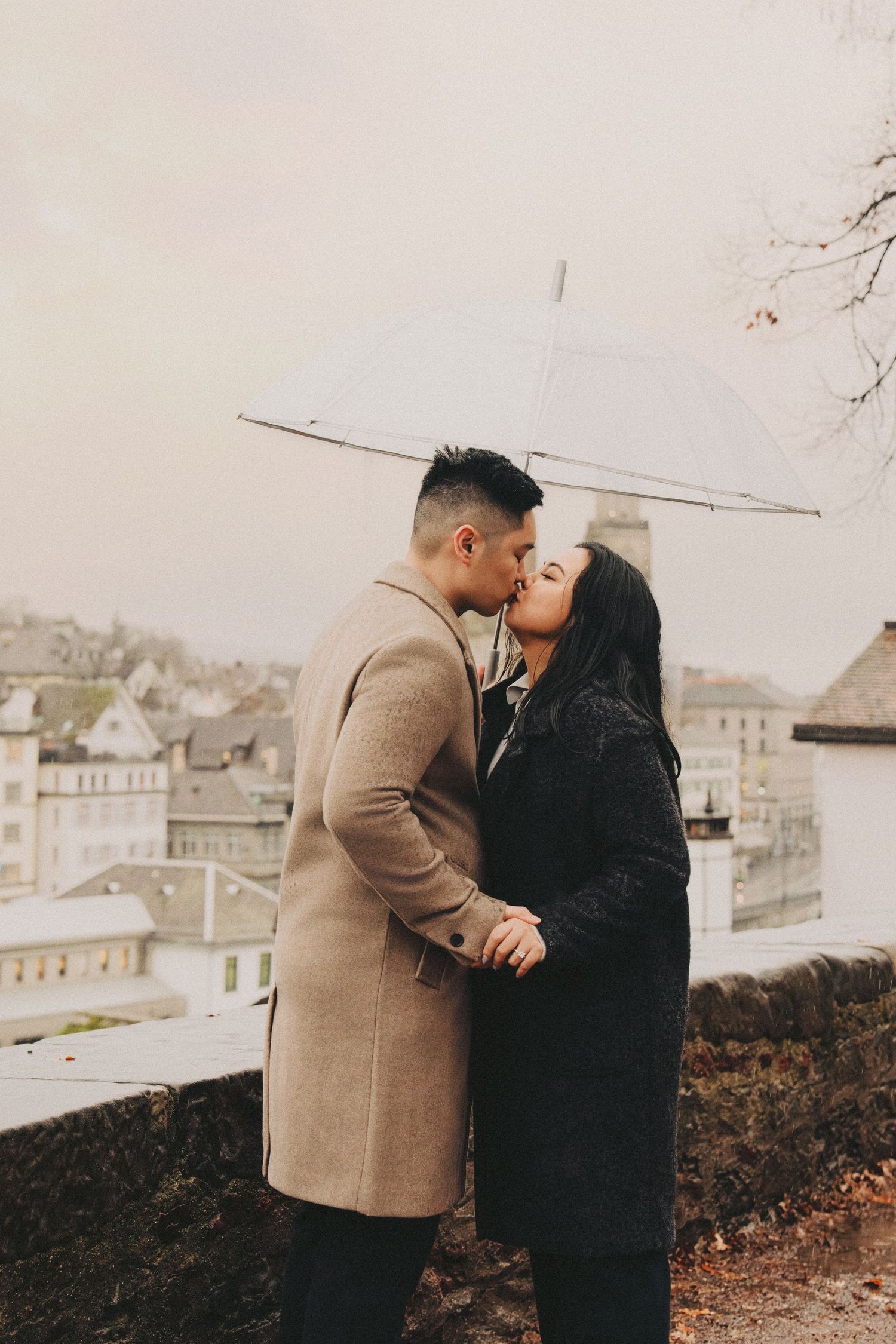 Couple kissing under clear umbrella on rainy day with European city rooftops in background.