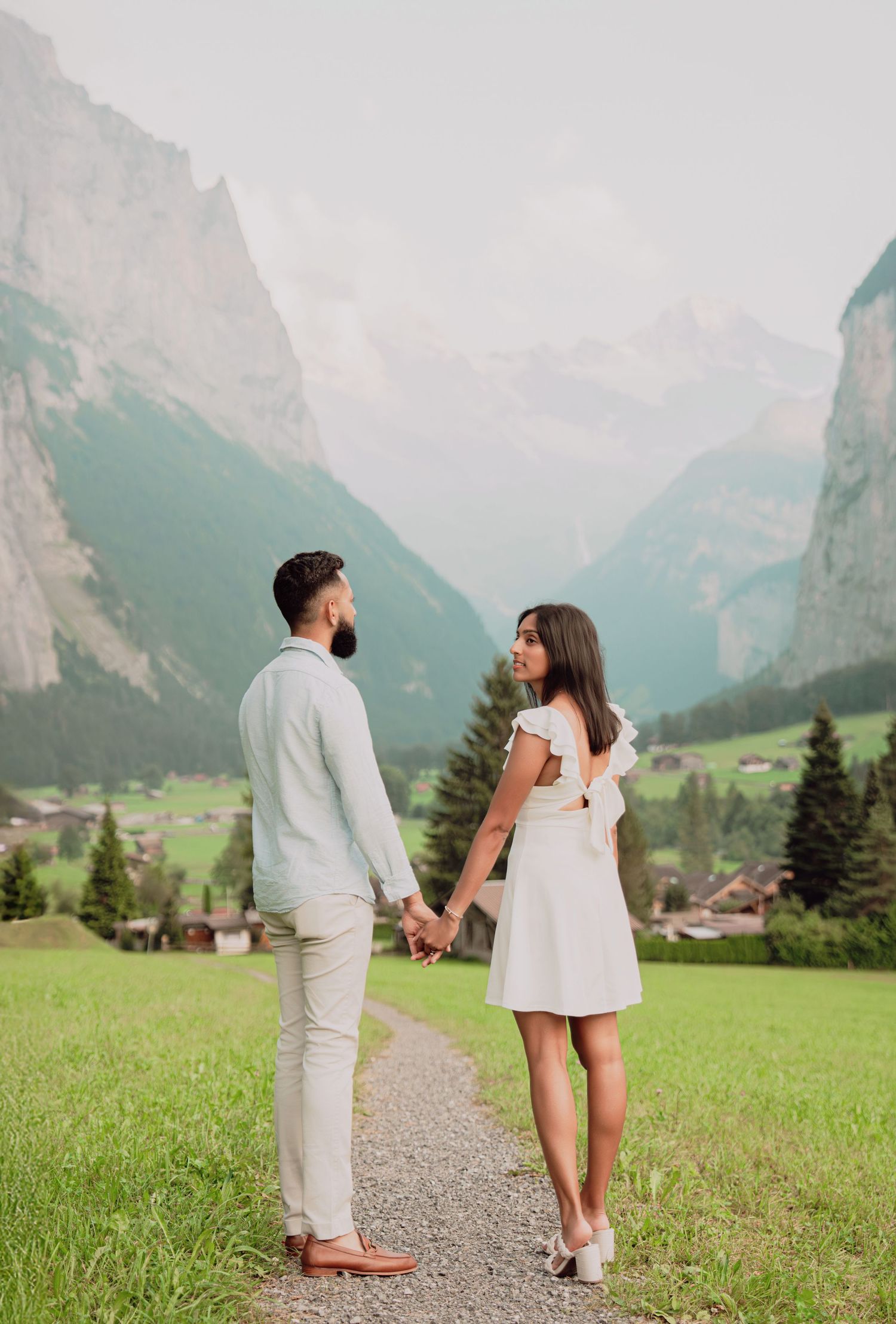 Couple holding hands on alpine path with sweeping view of misty mountain valley and green Swiss landscape behind them.