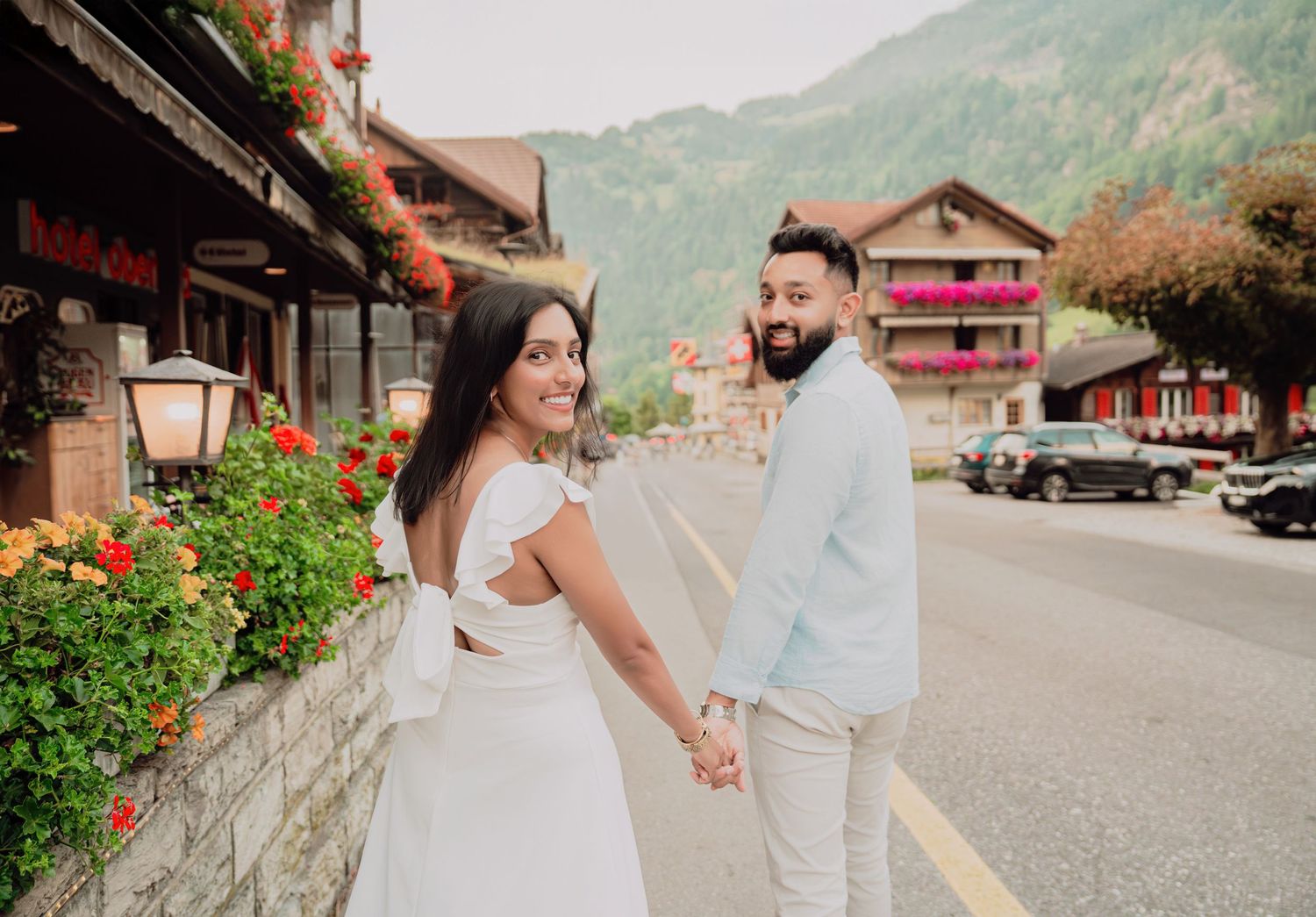 Couple holding hands on a flower-lined street in a Swiss alpine village, man looking back at camera.