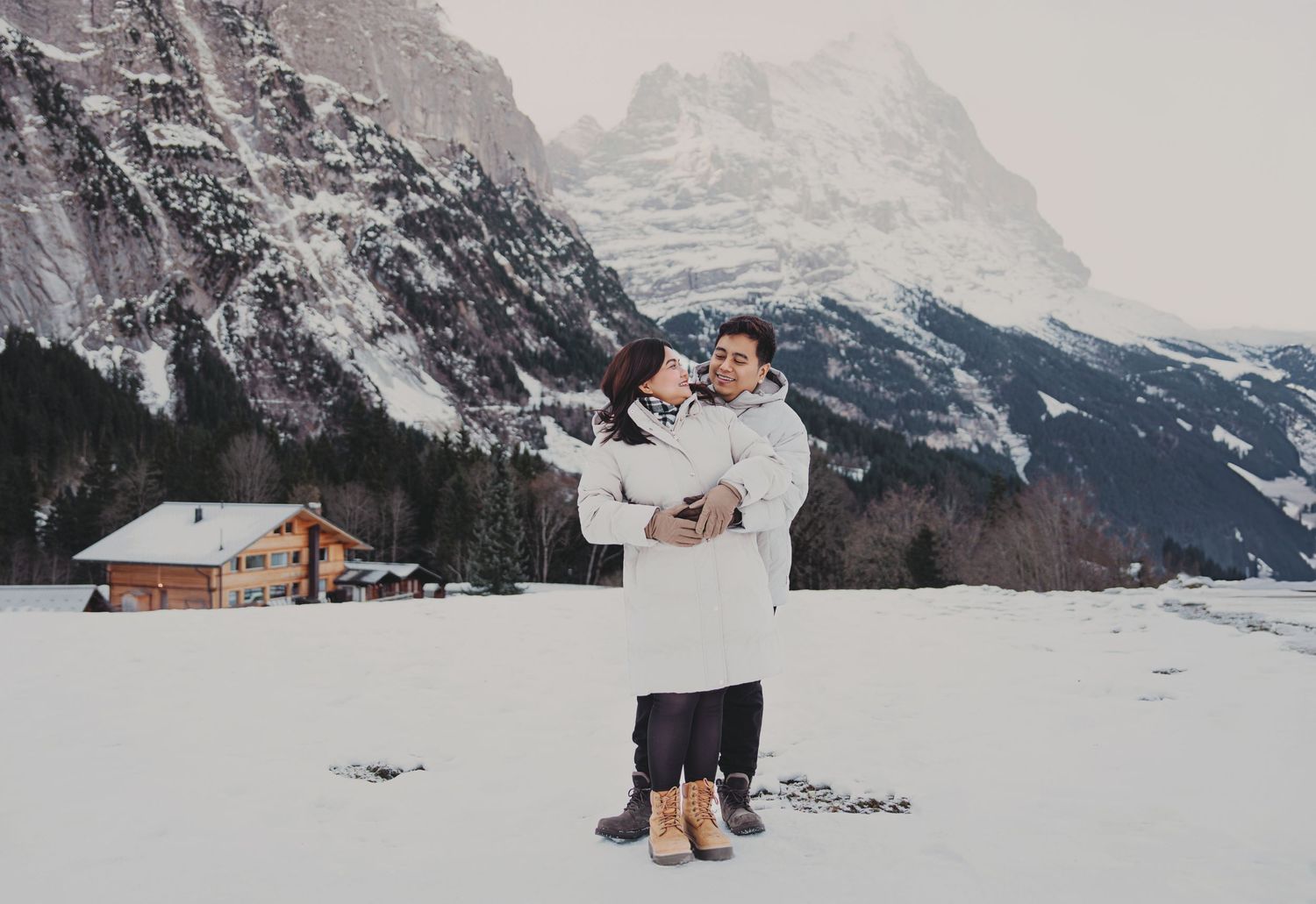 Couple embracing in snowy alpine landscape with mountain backdrop and wooden chalet in winter.