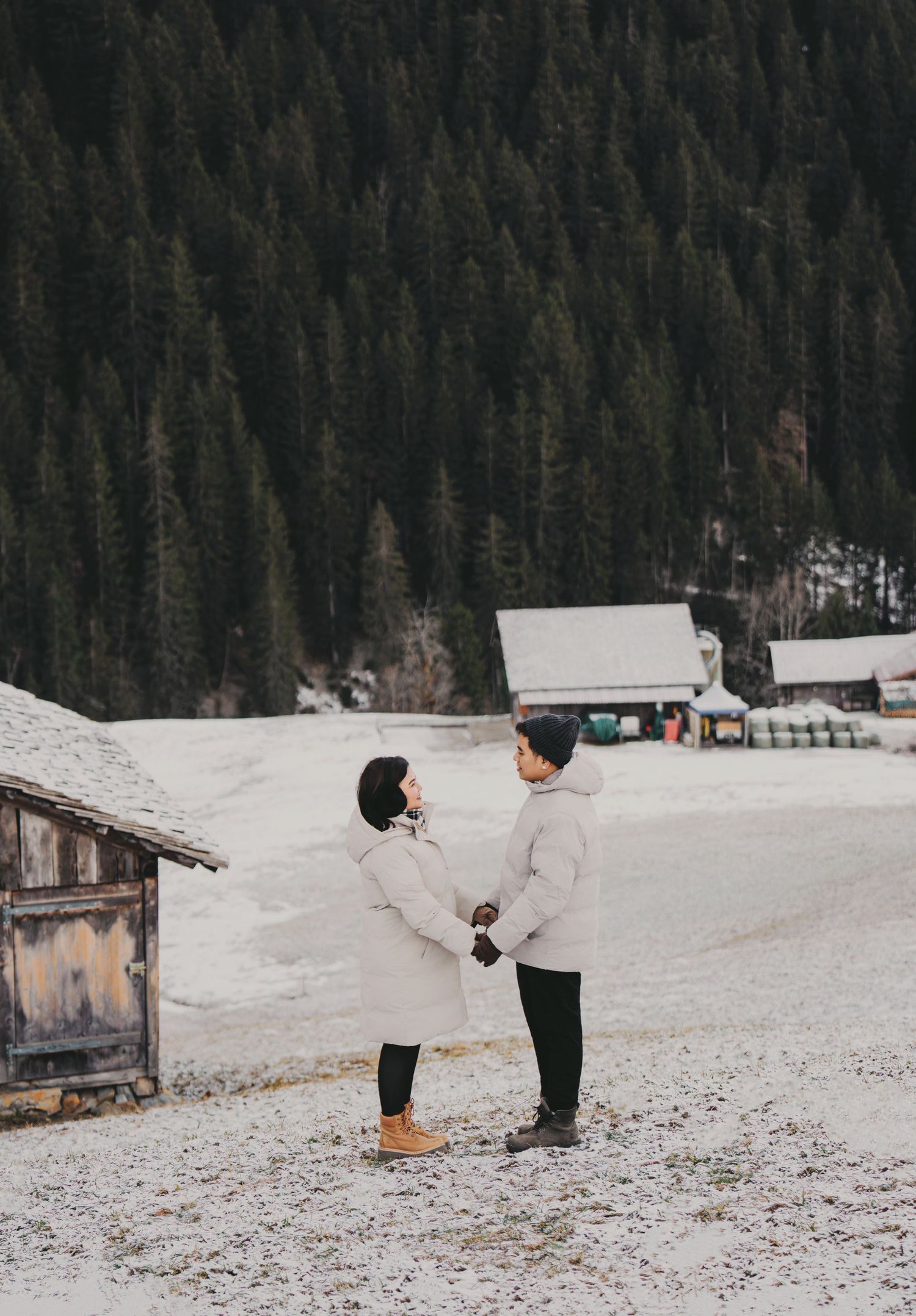 Couple holding hands in snowy alpine landscape surrounded by dense evergreen forest.