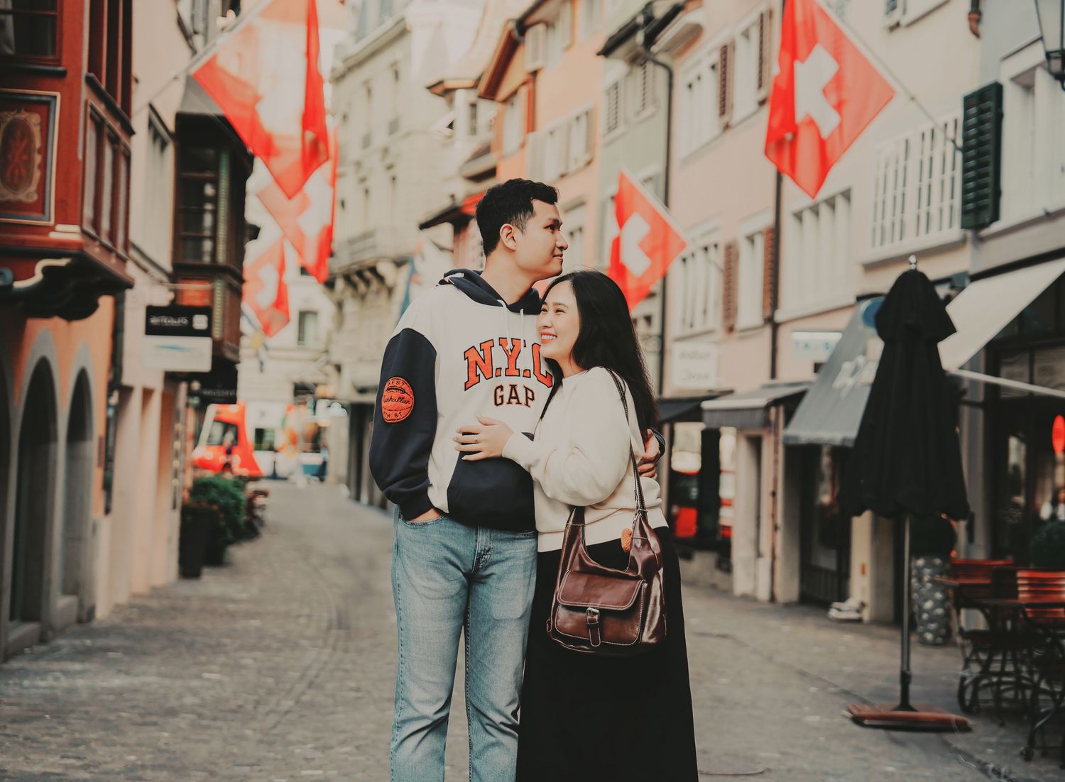 A couple shares a tender moment embracing on a colorful Swiss street lined with national flags.