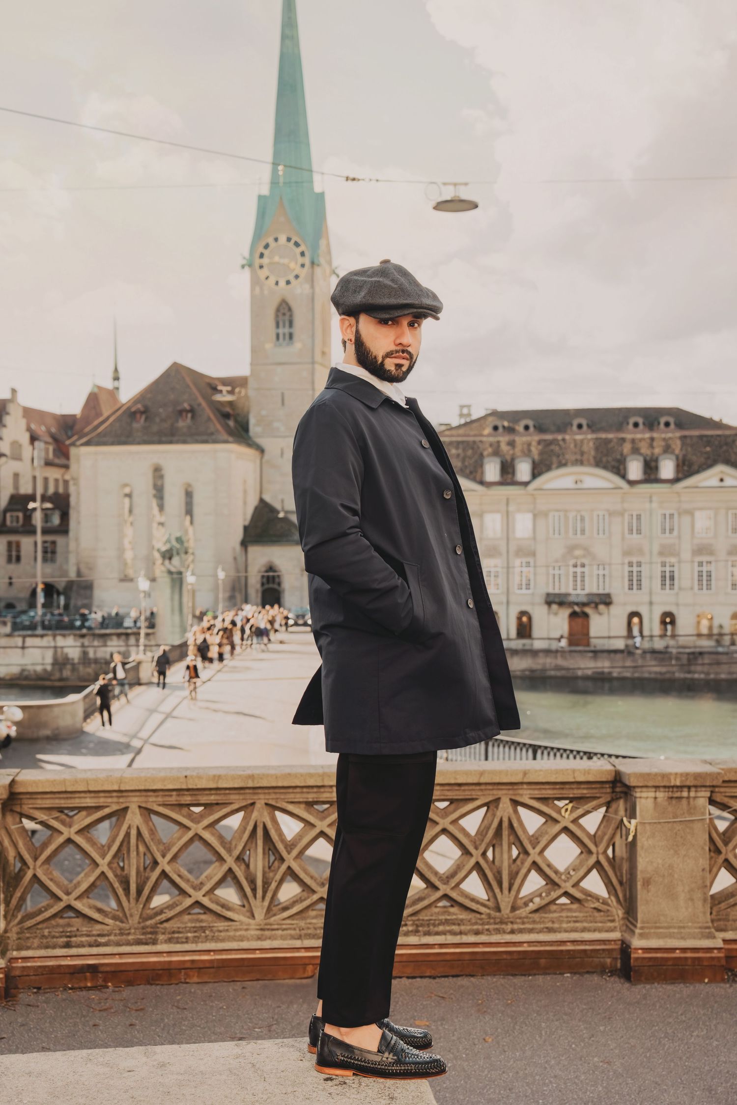 Dapper man in black coat and newsboy cap poses on Zurich bridge with historic church spire in background.