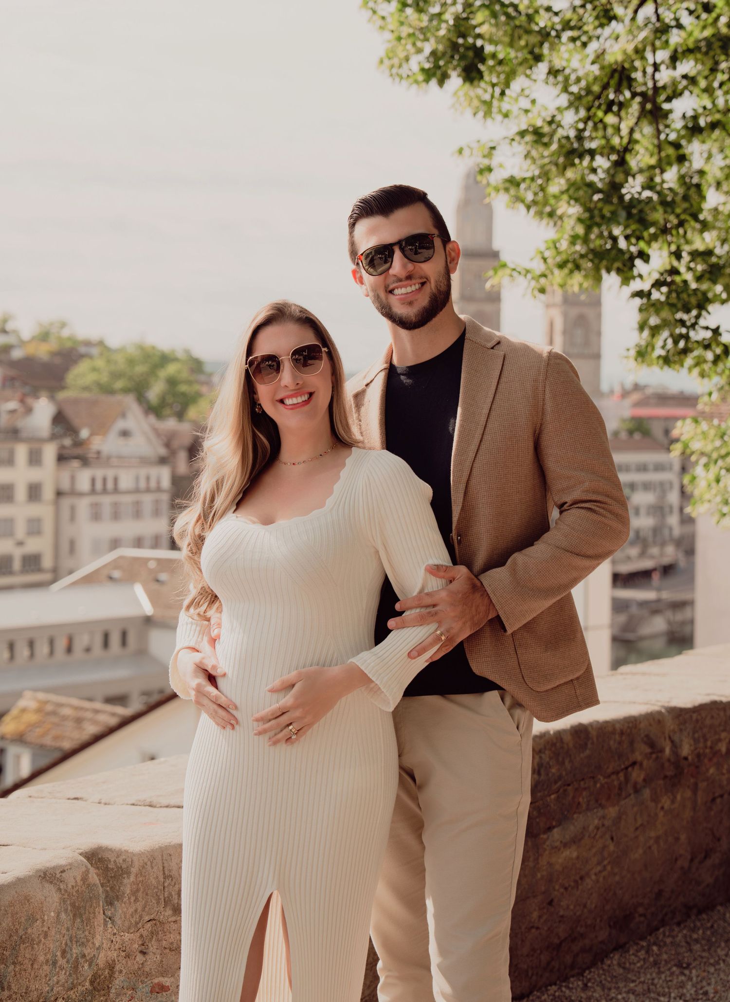 Smiling pregnant couple in sunglasses poses together on Zurich terrace with historic city skyline backdrop.