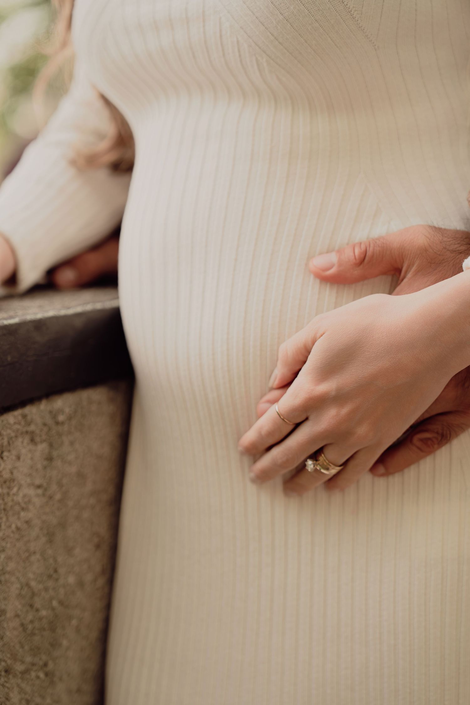 Close-up of pregnant womans belly with both partners hands gently resting on cream ribbed dress.