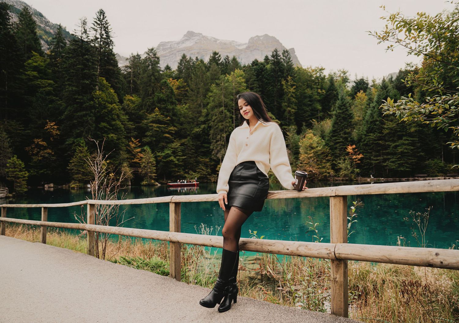 Young woman in cream sweater and black leather skirt posing by wooden fence at scenic turquoise mountain lake.