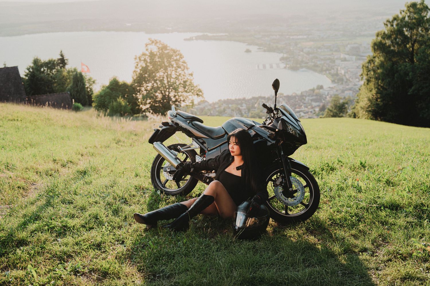 Young woman in black outfit relaxing beside a black sport motorcycle on a hillside with lake view at sunset.