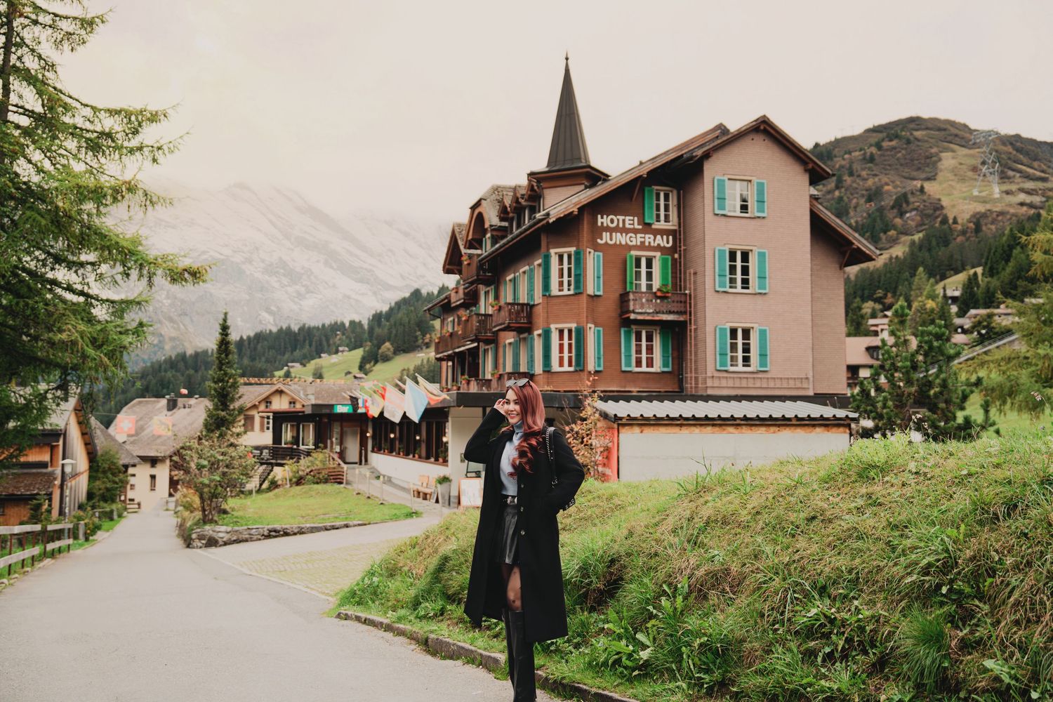 Woman in long black coat walks past Hotel Jungfrau amid lush green Swiss mountain landscape.
