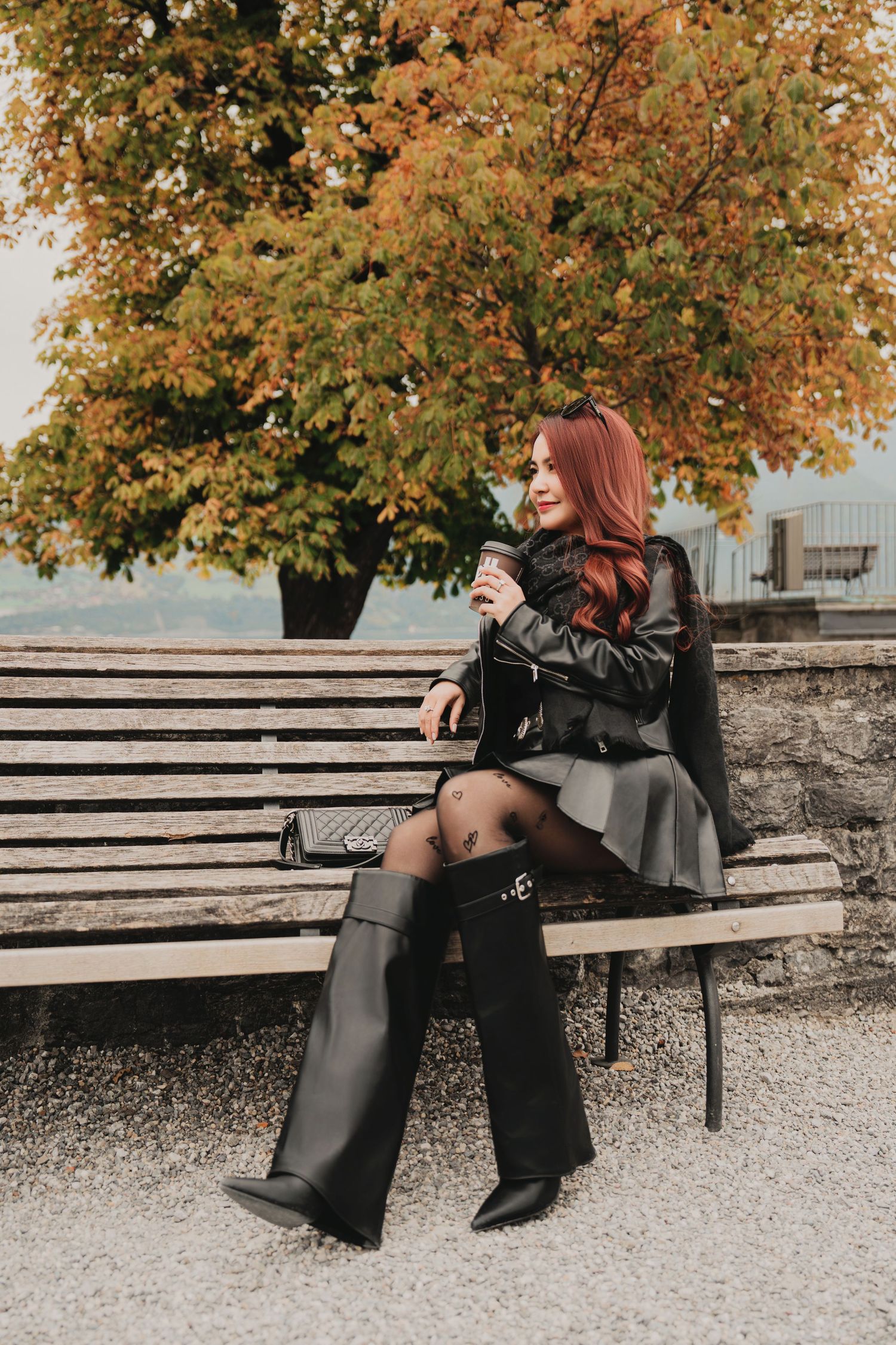 Stylish woman with red hair holding coffee on a wooden bench surrounded by fall foliage.