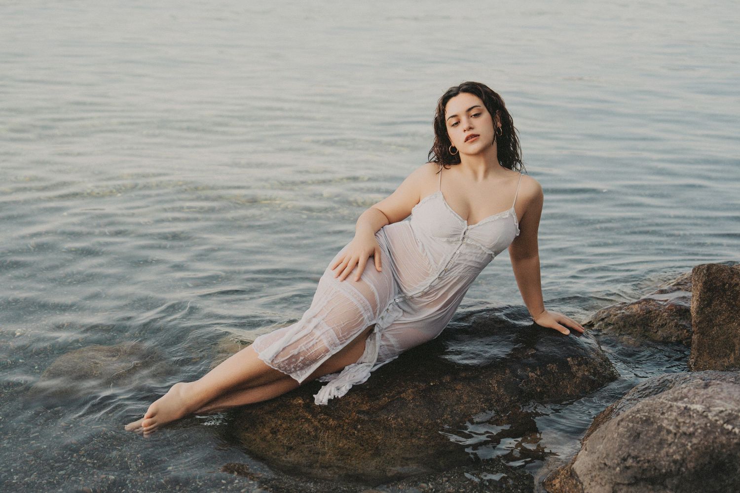 Woman in wet white lace dress reclining on rocks at waters edge.