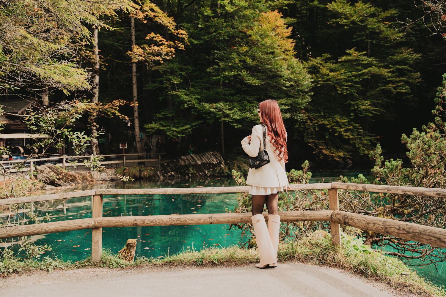Woman with long red hair in white outfit gazing at turquoise lake in autumn forest.