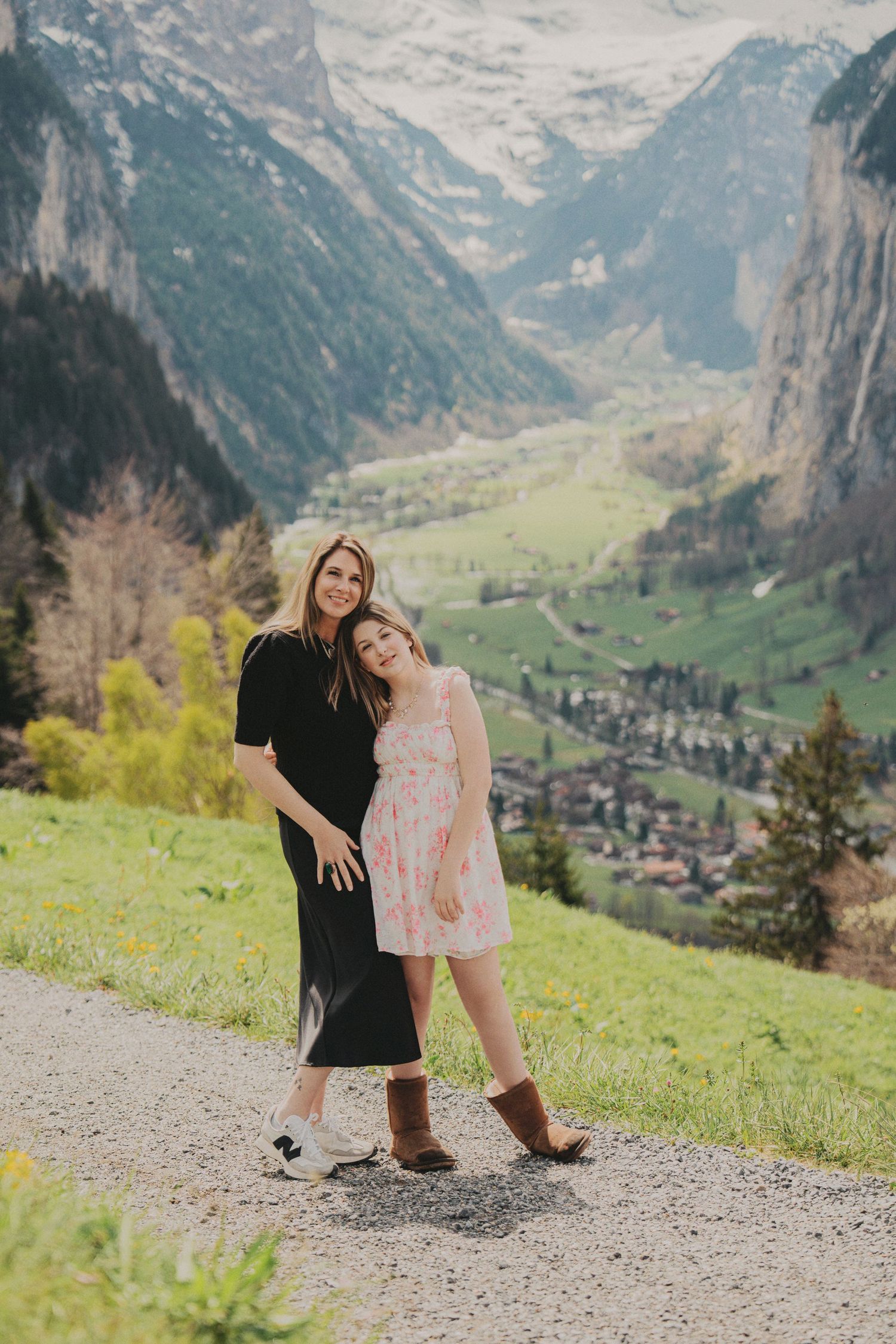Woman in black dress and girl in pink floral dress pose together on mountain path in Swiss Alps valley.