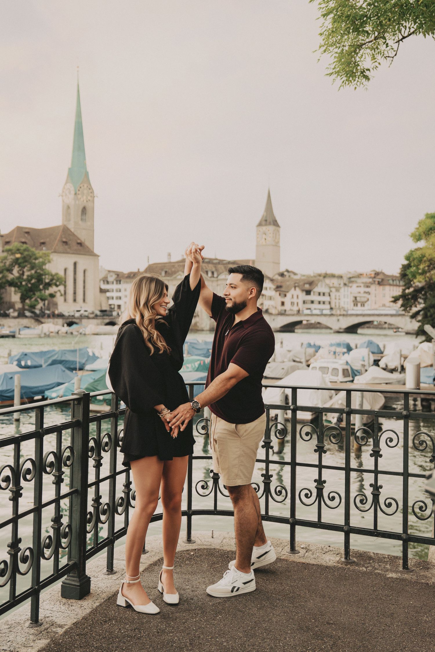 Couple holding hands and dancing on a balcony with Zurich church spires and river in background.