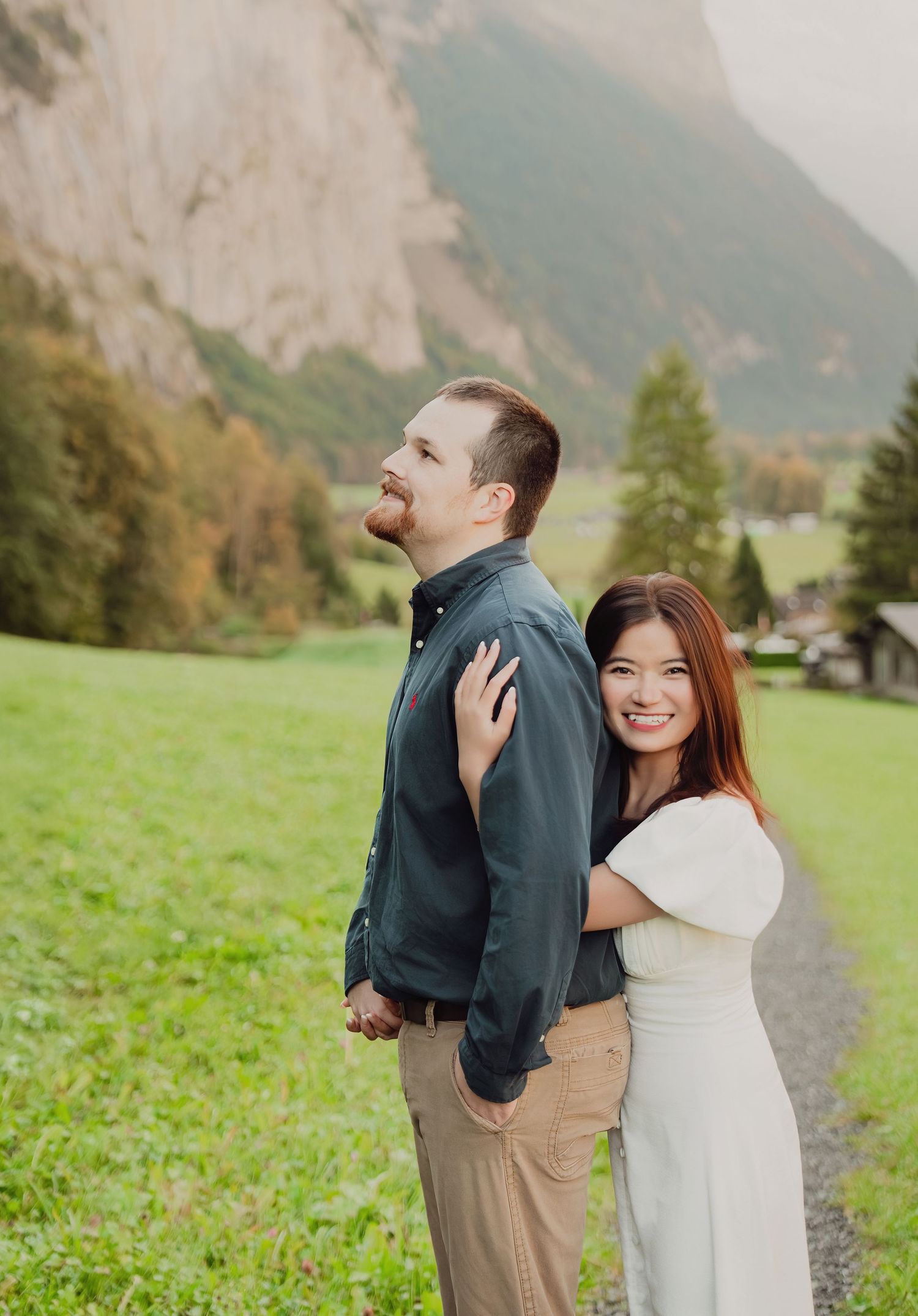 Smiling woman embracing man from behind on a path with green alpine landscape.