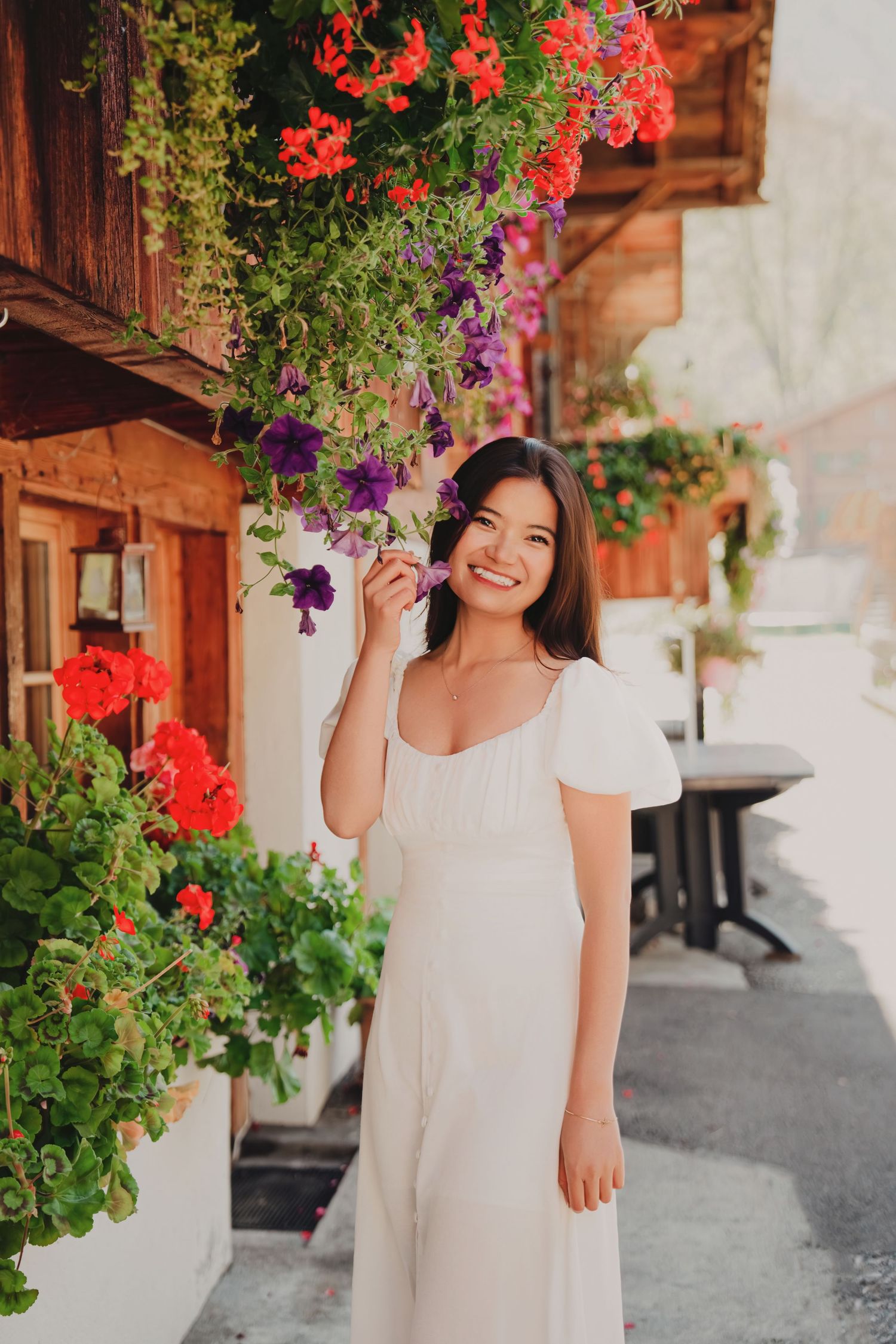 Smiling young woman in a white dress holding purple petunias beside colorful hanging flower baskets on a wooden building.