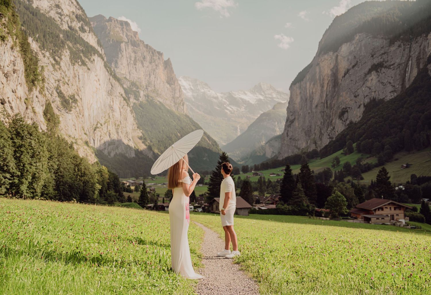 Woman in white dress holding parasol facing man on a scenic path in Lauterbrunnen, Switzerland.