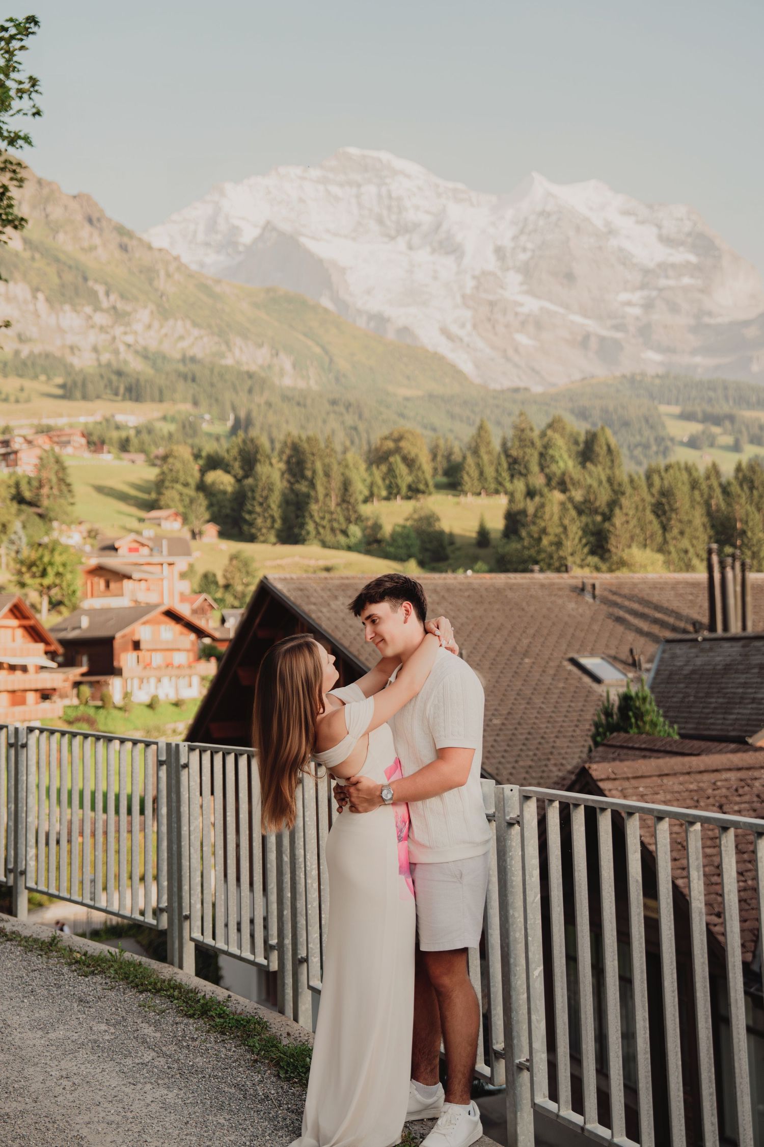 Romantic couple hugging on a hillside path overlooking charming Swiss chalets and glacial mountain scenery.