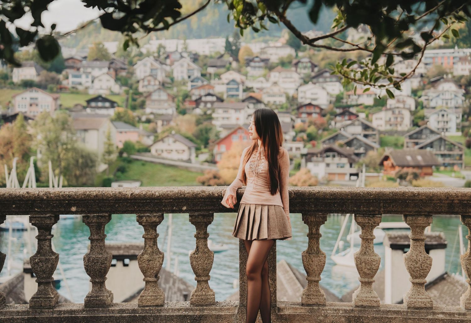 Young woman in a skirt stands at a stone balustrade overlooking a scenic Swiss lakeside village.
