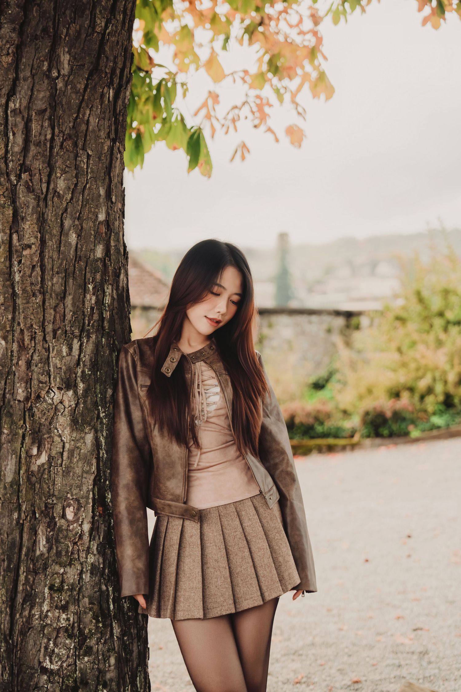 Smiling young woman in a brown jacket and pleated skirt leaning against a tree in an autumn park setting.