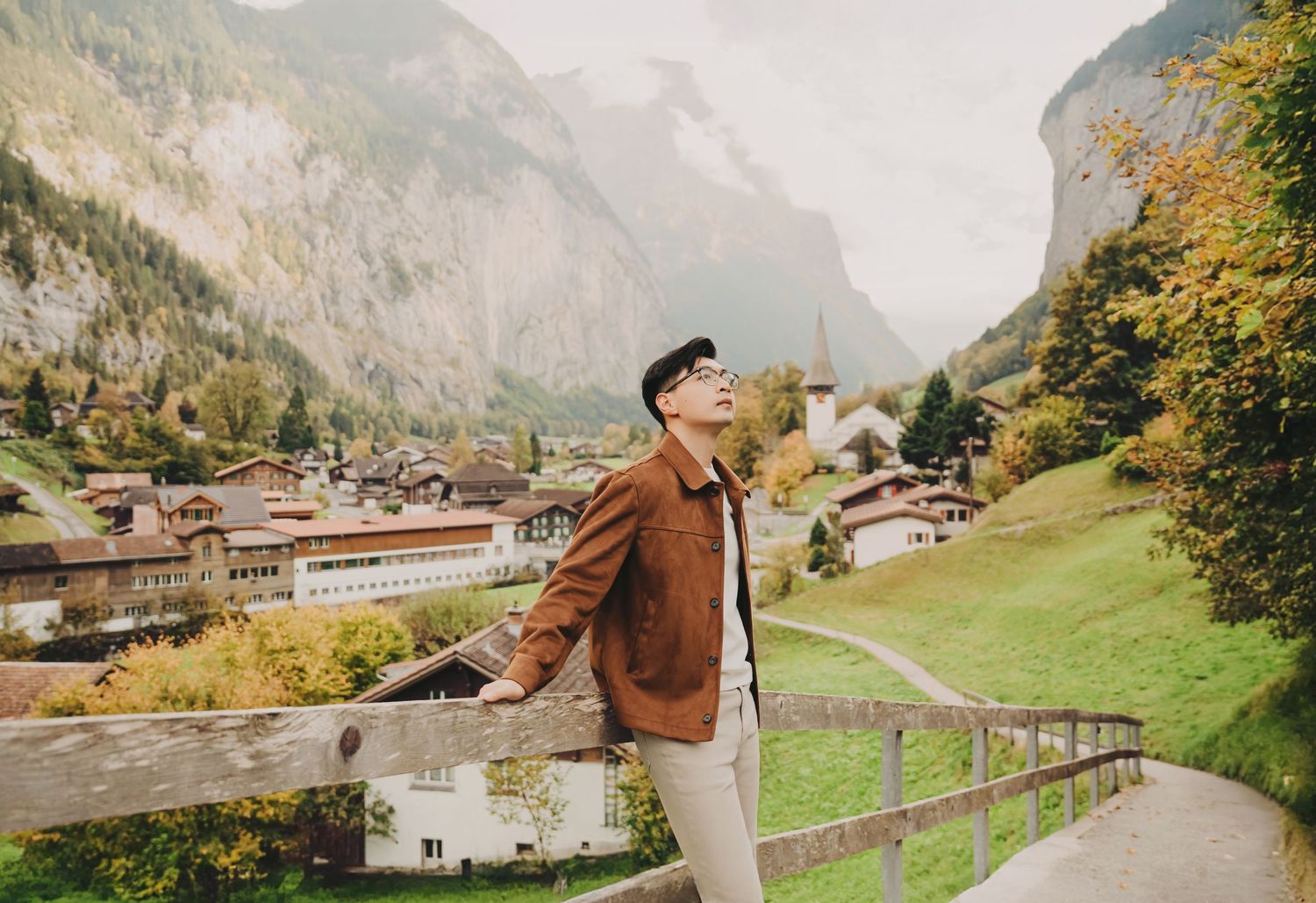 Man in brown jacket poses by fence with stunning Swiss alpine village and rocky mountain cliffs behind.