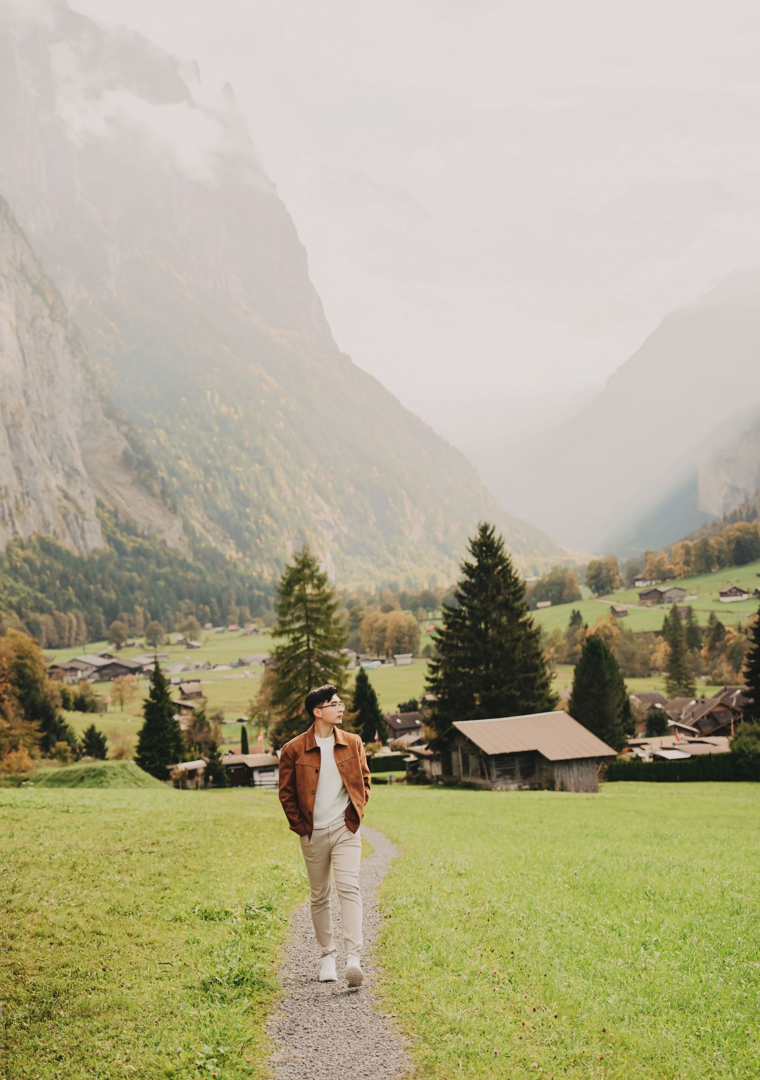 Man in brown jacket walking on a path through a scenic Swiss alpine valley with misty mountains.