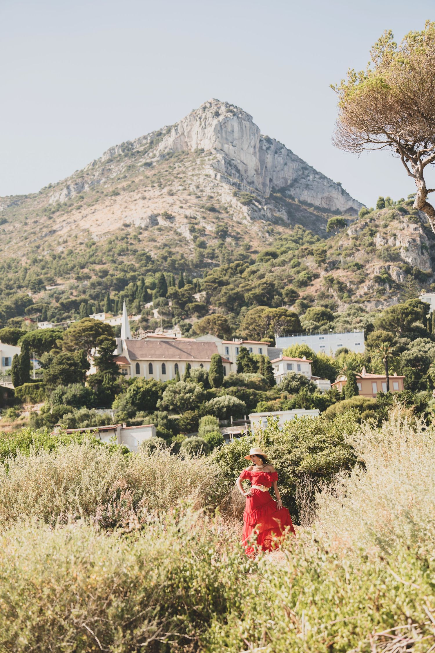 Woman in red dress standing in Mediterranean hillside landscape with mountain and village backdrop.