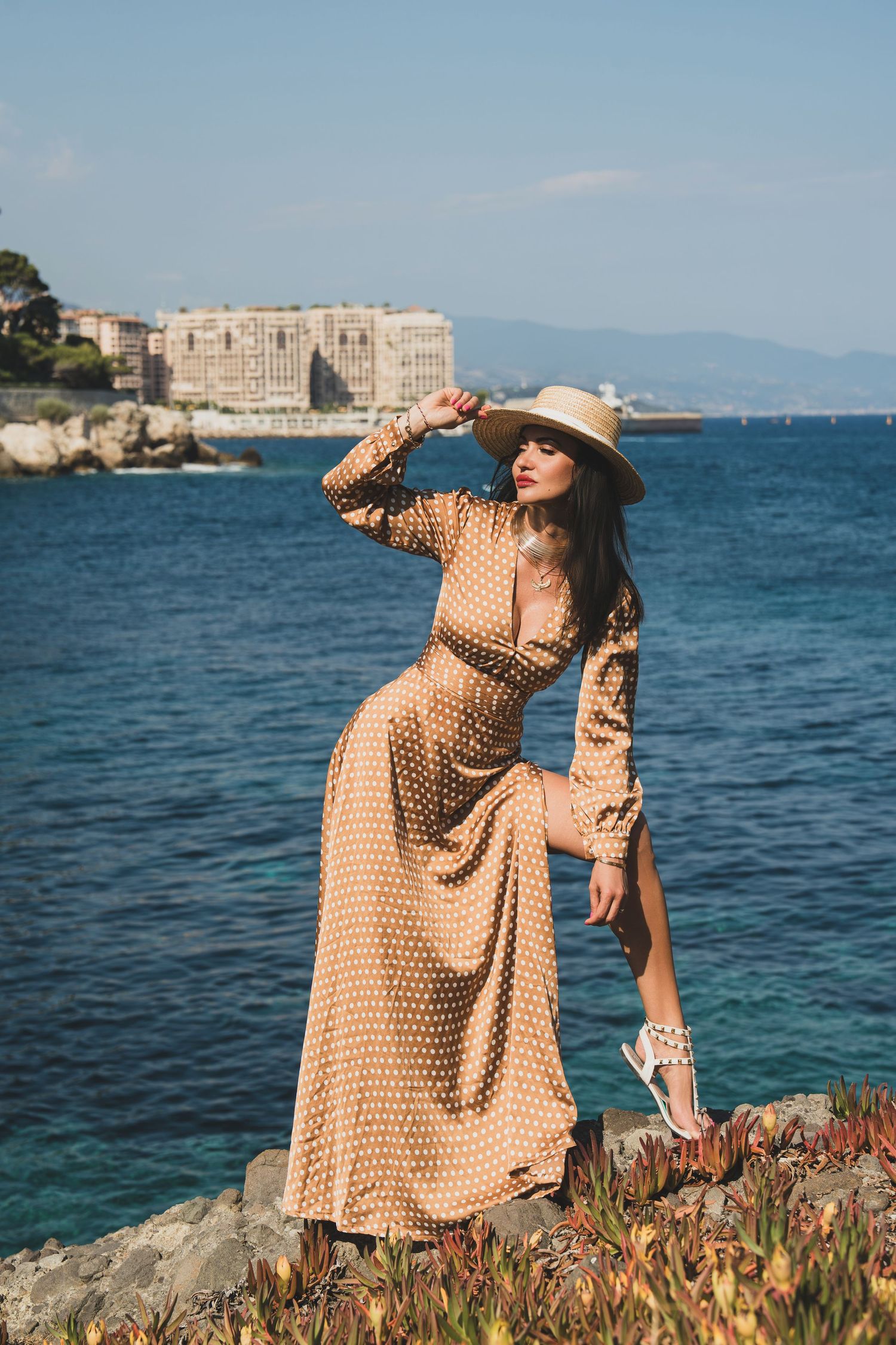 Woman in gold polka dot maxi dress and straw hat posing on rocky Mediterranean coastal scenery.