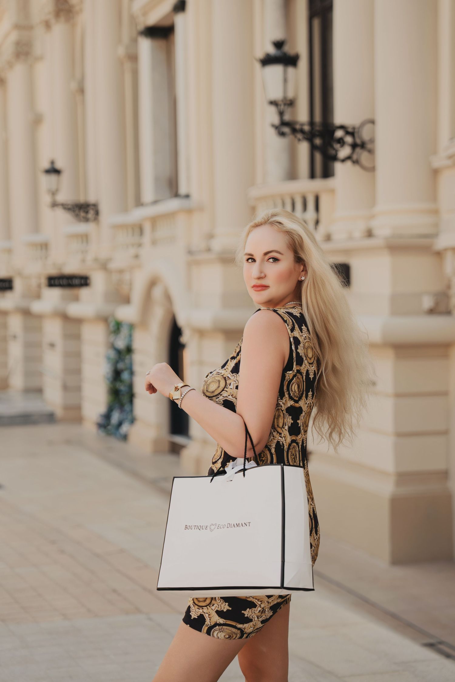 Stylish blonde woman in Versace-print dress carrying white luxury shopping bag outdoors.