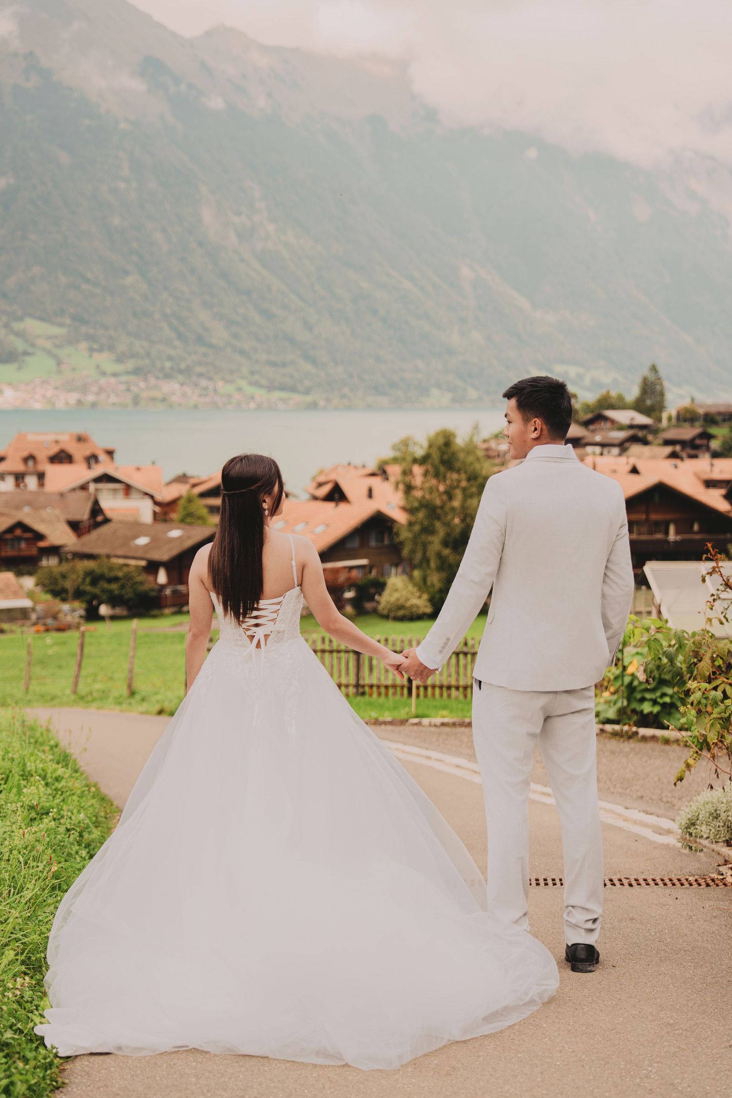 Bride and groom walking hand in hand toward scenic Swiss lakeside village with alpine mountains.