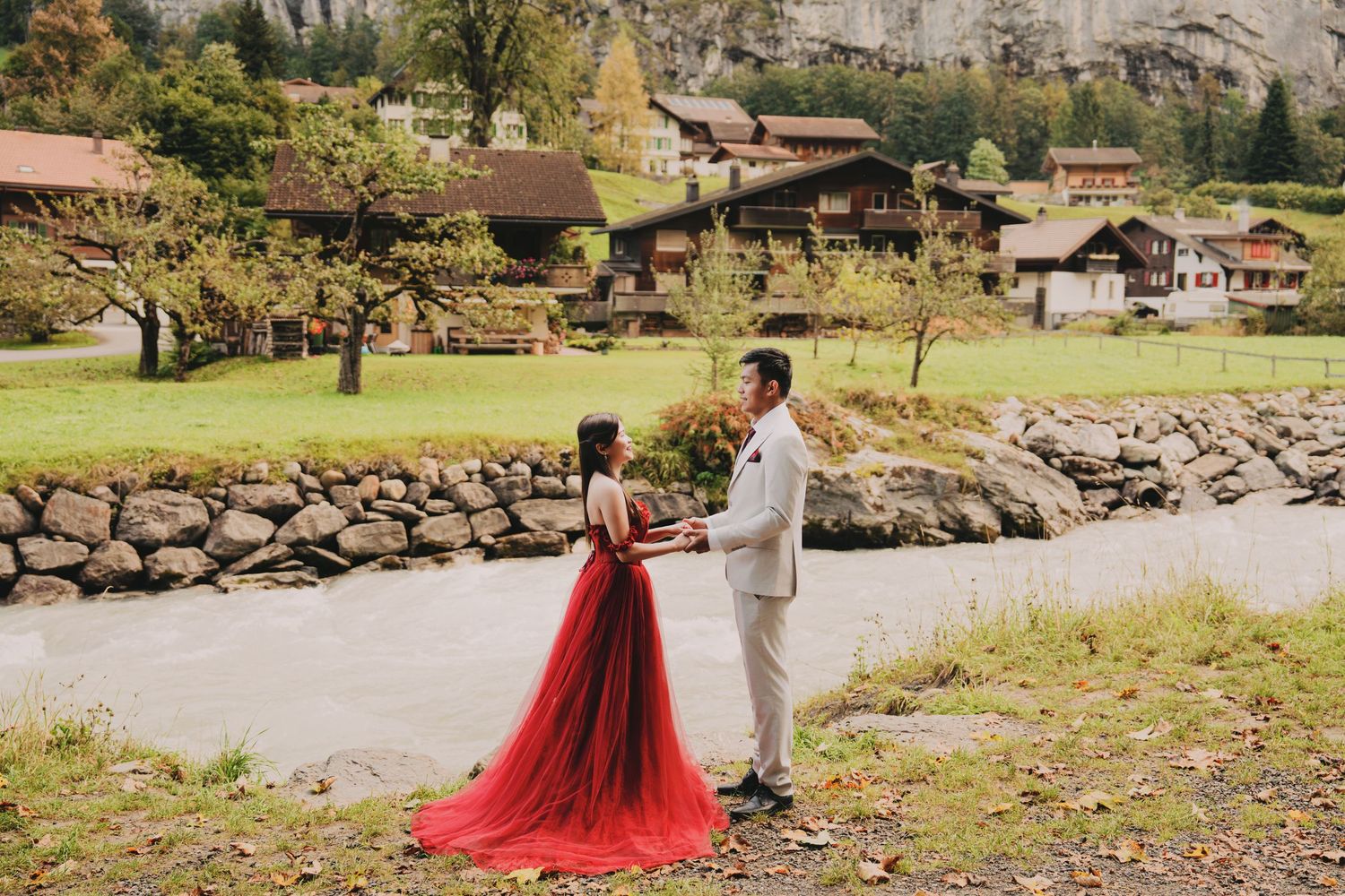 Pre-wedding couple facing each other by stone wall river in picturesque Swiss alpine village setting.