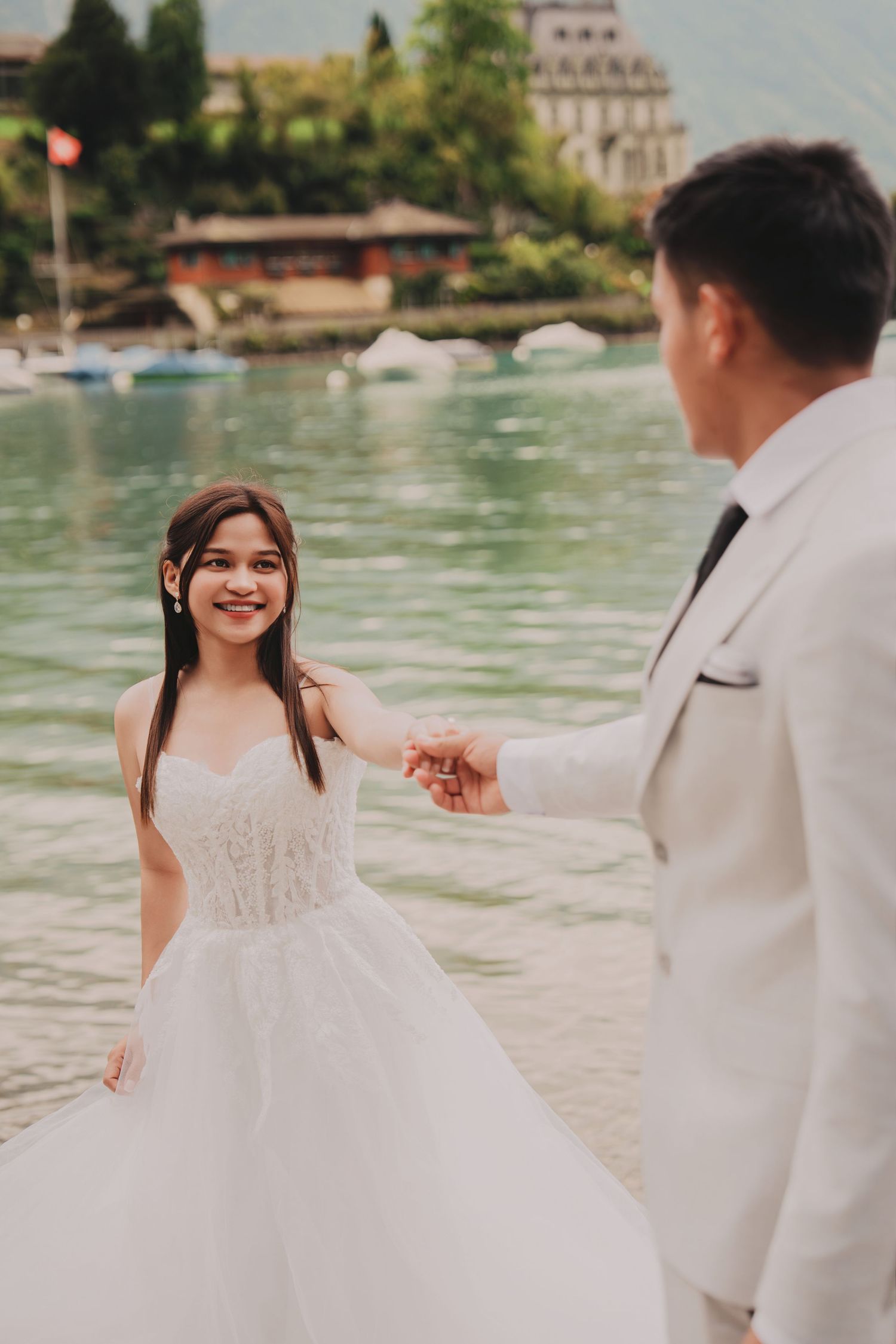 Bride in strapless lace gown smiling while holding groom's hand by a picturesque alpine lakeside.