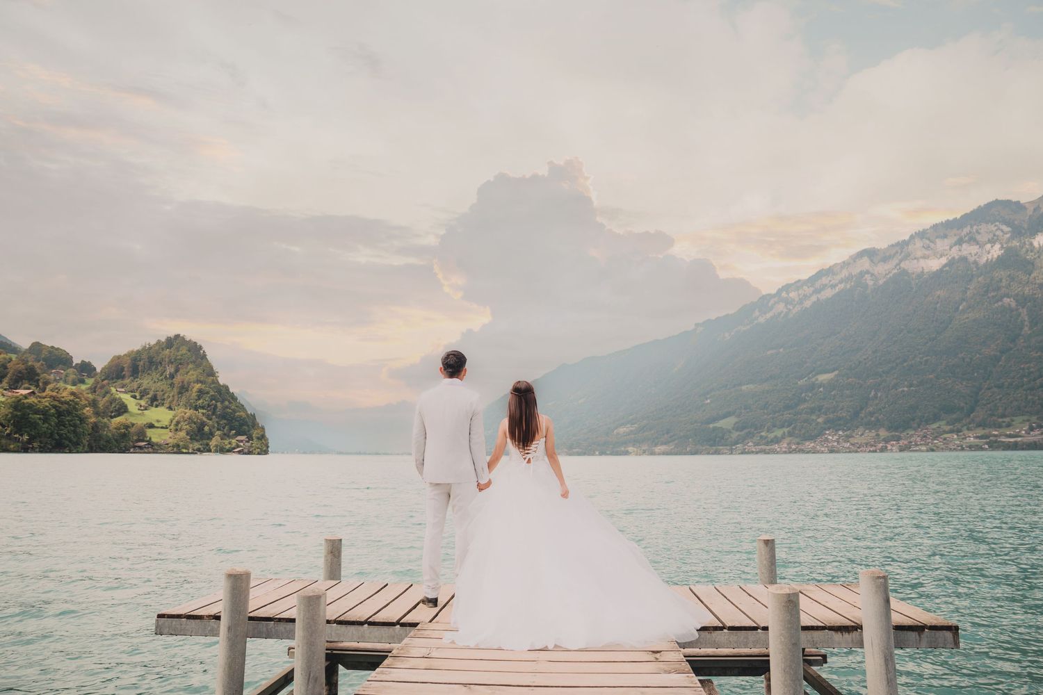 Wedding couple holds hands walking on wooden dock overlooking stunning alpine lake and mountain sunset scenery.