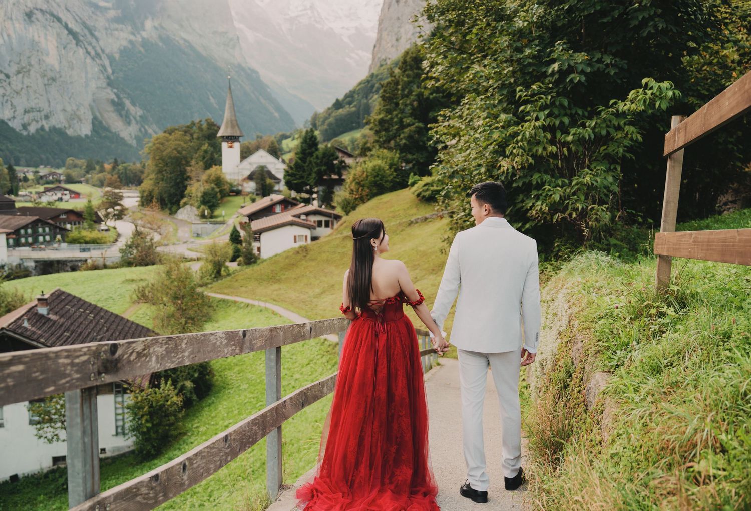 Couple strolling hand-in-hand toward picturesque Swiss alpine village with church and mountain views.