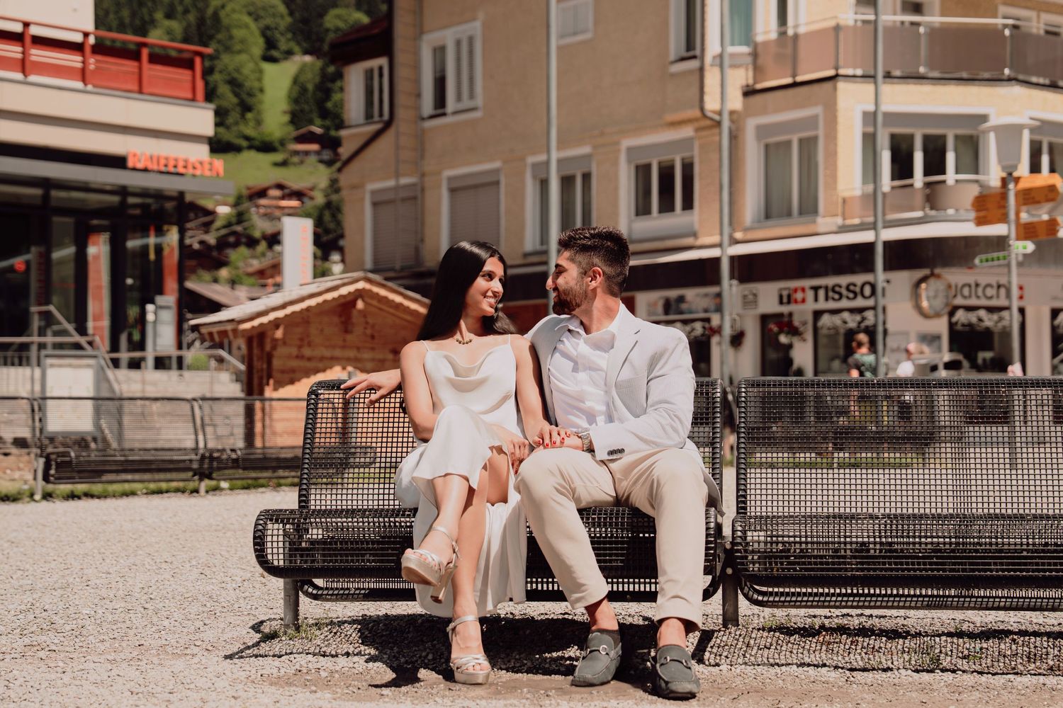 Happy couple sitting on a bench chatting in a sunny Swiss village square on a bright day.