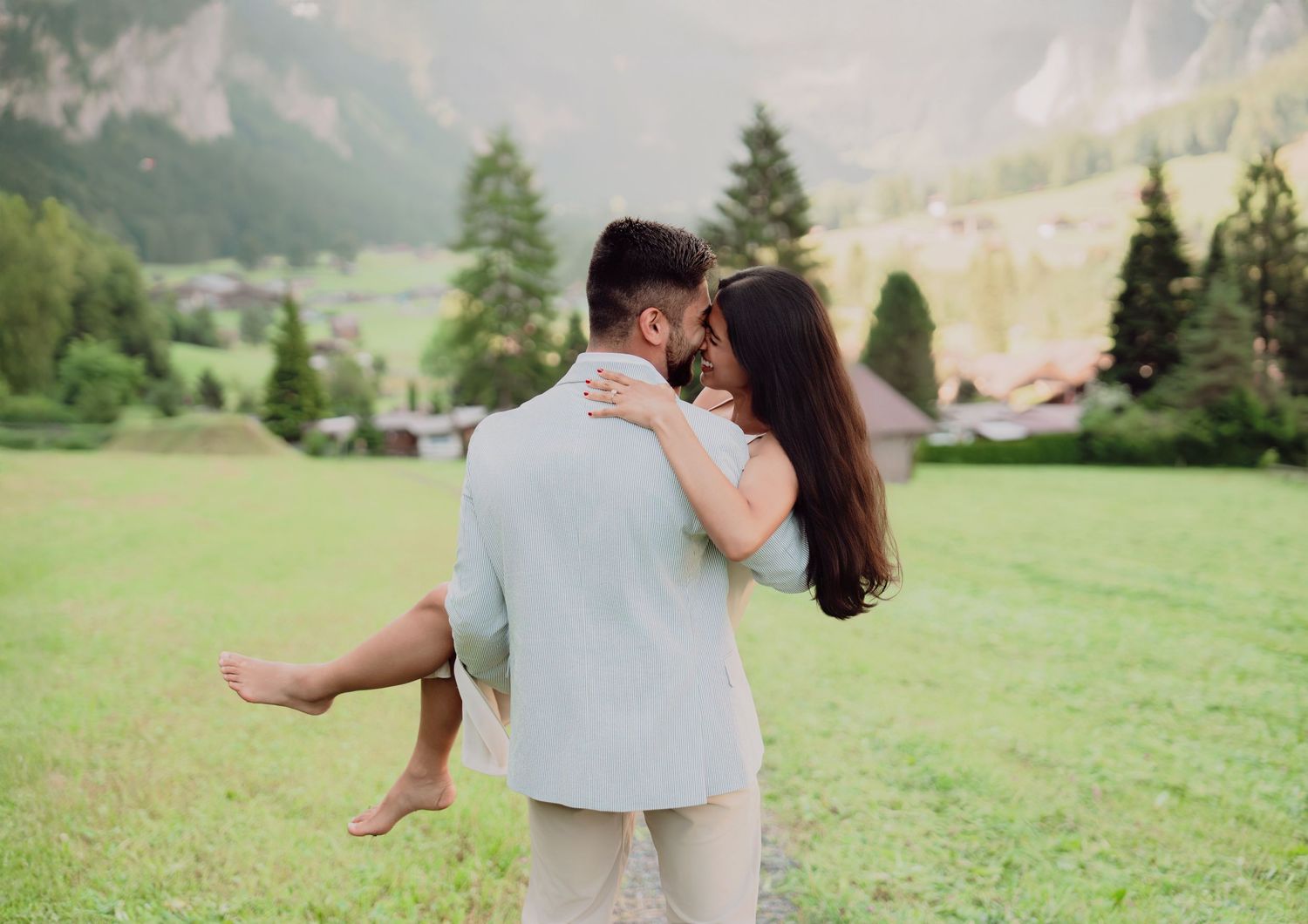 Man lifting woman in his arms in a green alpine meadow with Swiss mountain village behind them.