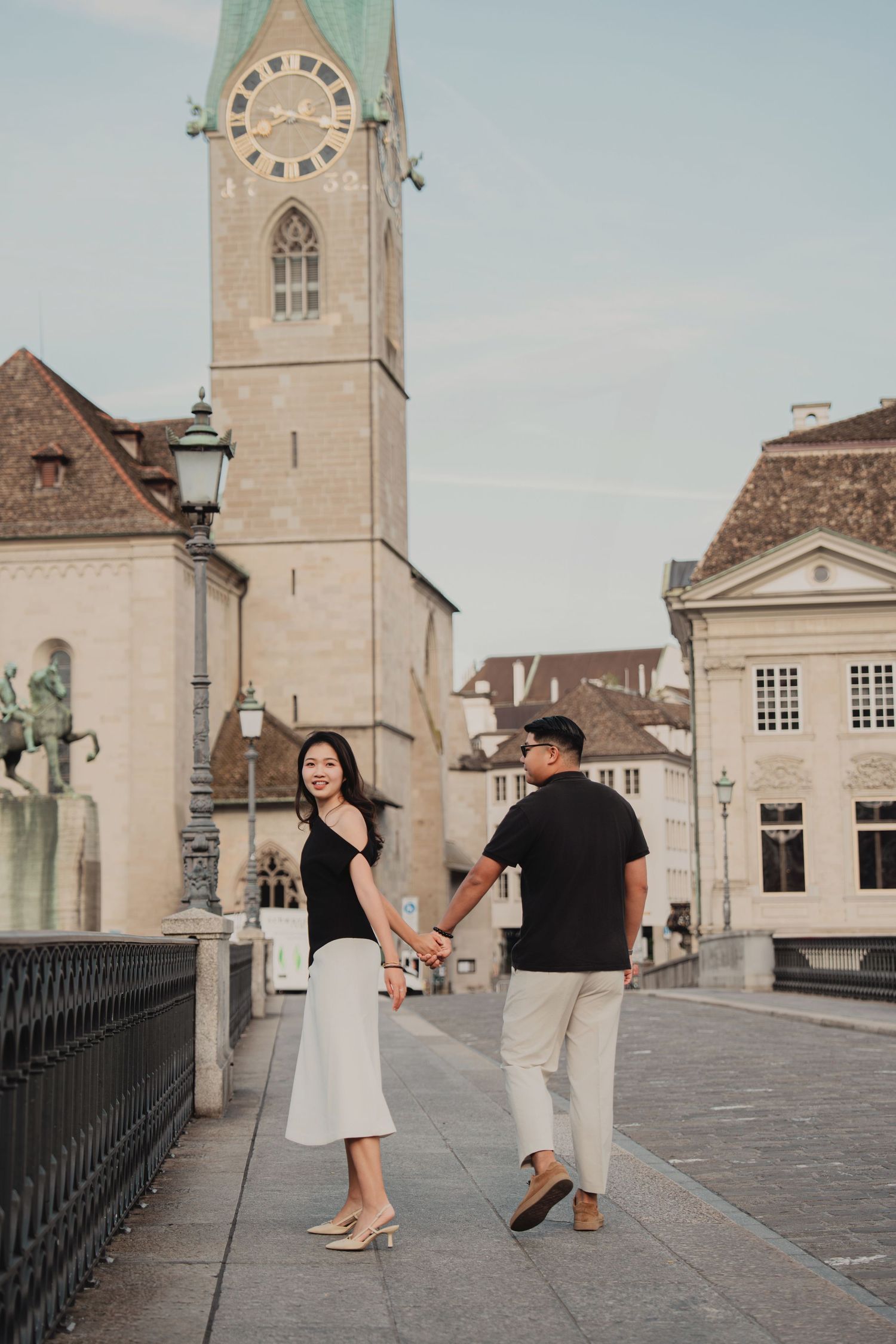 Woman smiling at partner while holding hands on a Zurich bridge with Fraumunster church tower.