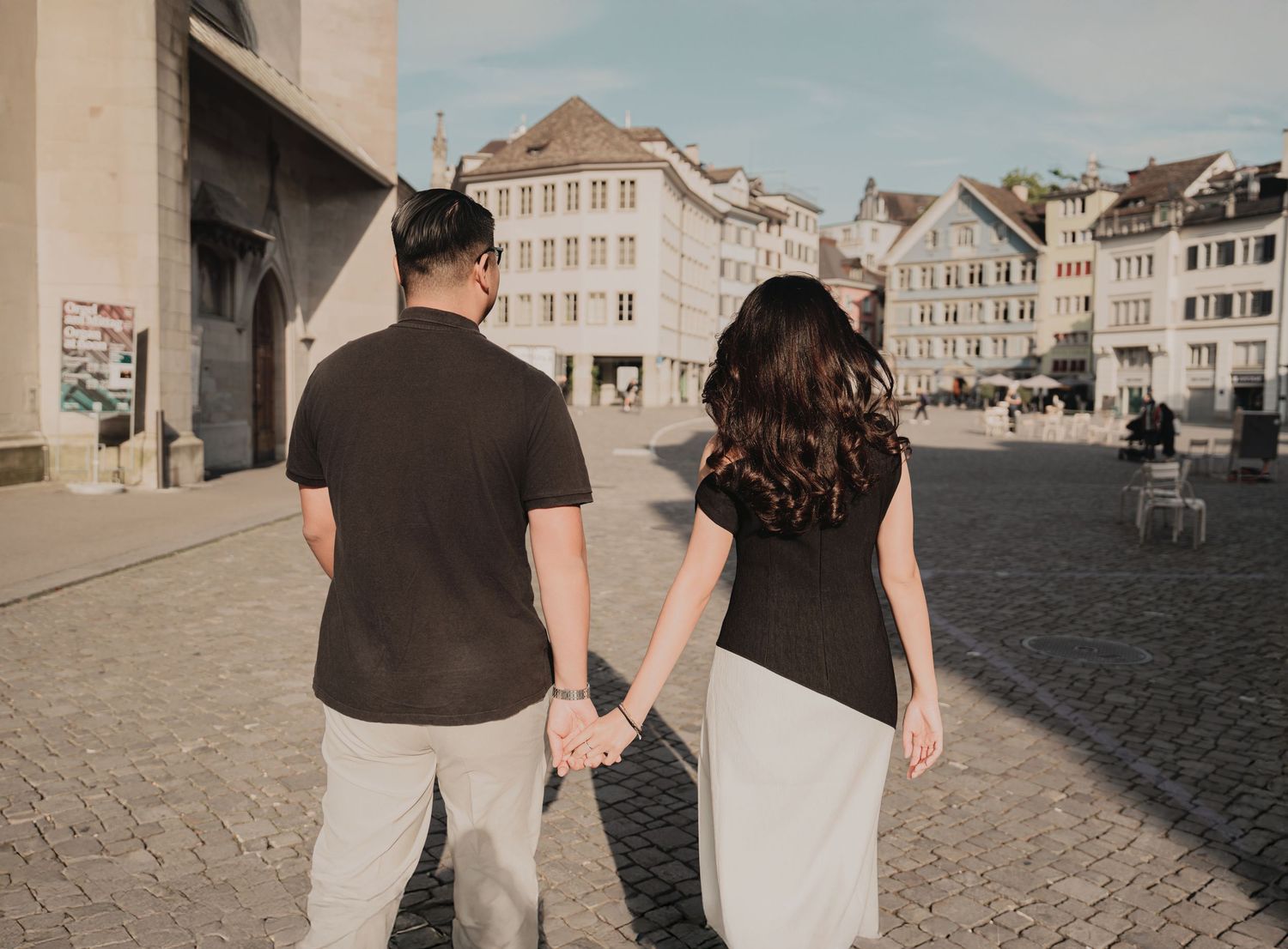 Couple holding hands walking through a European cobblestone square, both dressed in black tops and light bottoms.