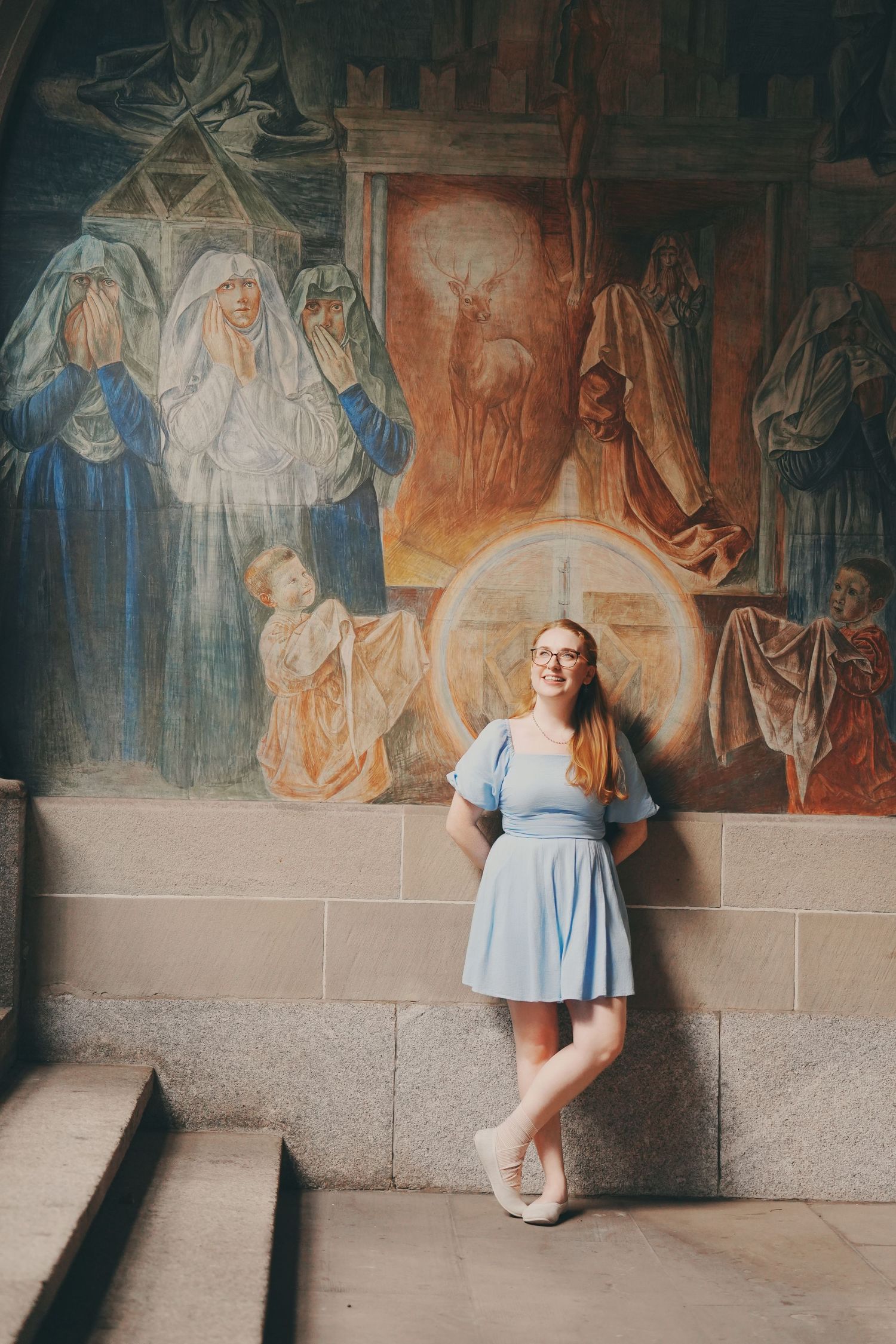 Smiling woman in blue dress leaning against wall with colorful religious fresco in background.