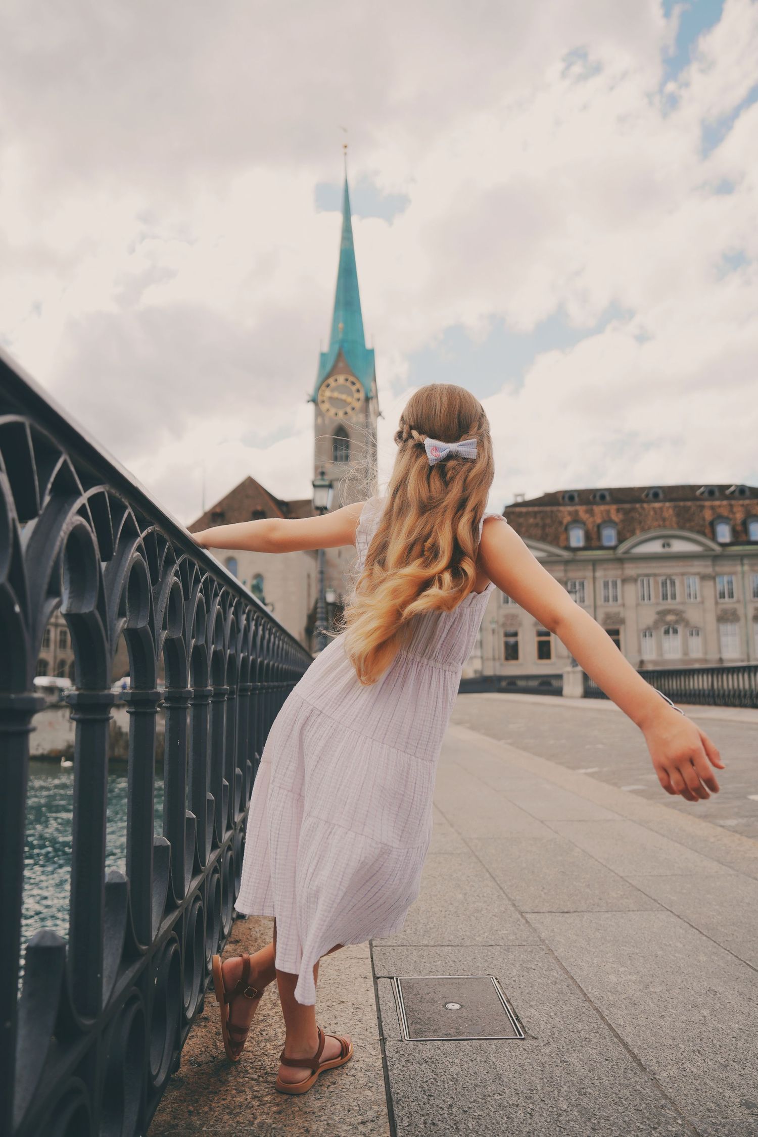 A young girl in a white dress walks along a bridge railing with Fraumunster church spire in Zurich, Switzerland.