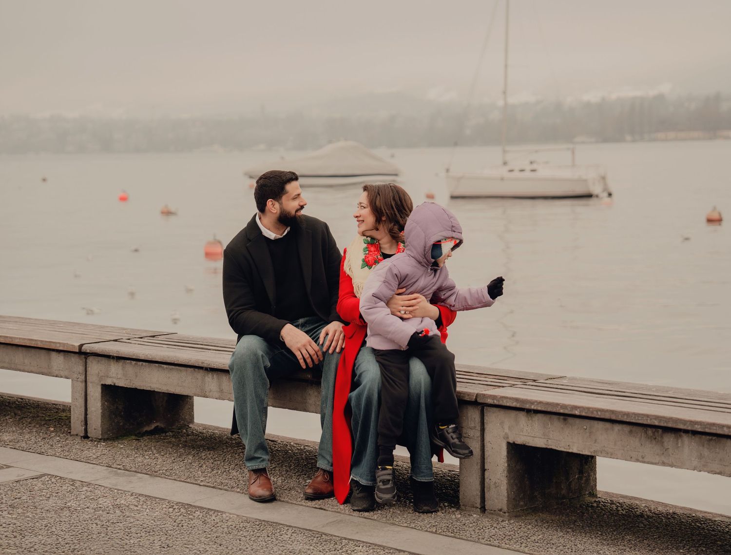 Family of three sits on a waterfront bench, parents chatting while holding their young child.