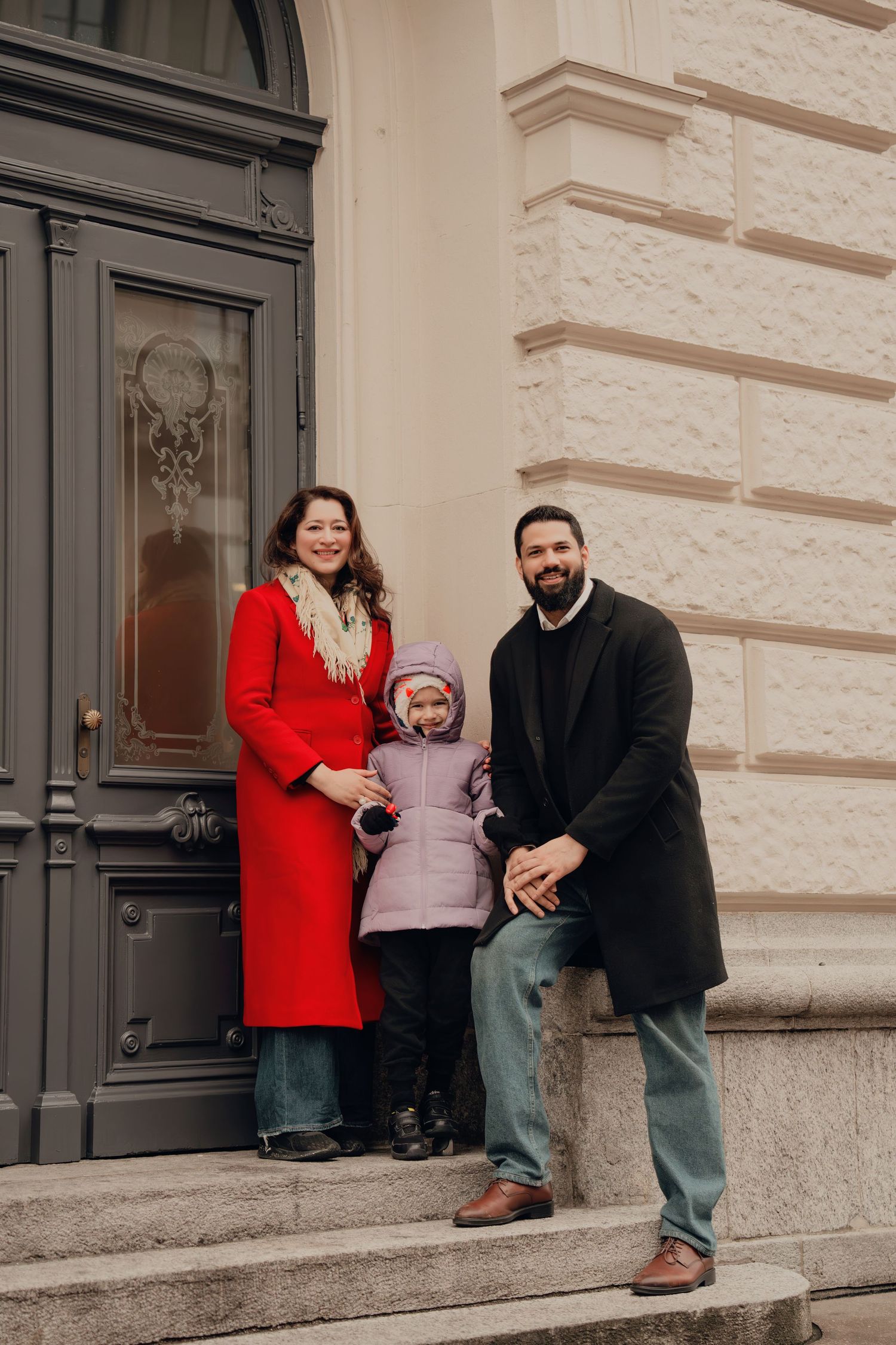Smiling family of three posing on steps before an ornate gray door of a classic building.