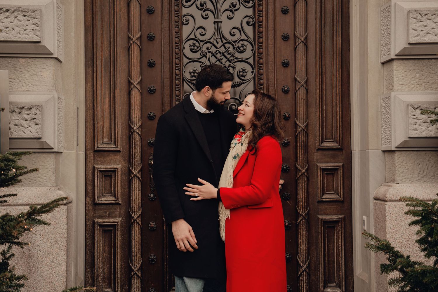 Romantic couple sharing a tender moment in front of an ornate wooden door, woman in red coat.