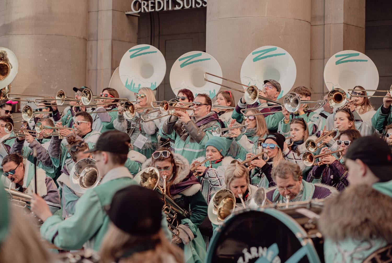 A large marching band in teal uniforms performs outdoors near a Credit Suisse building.