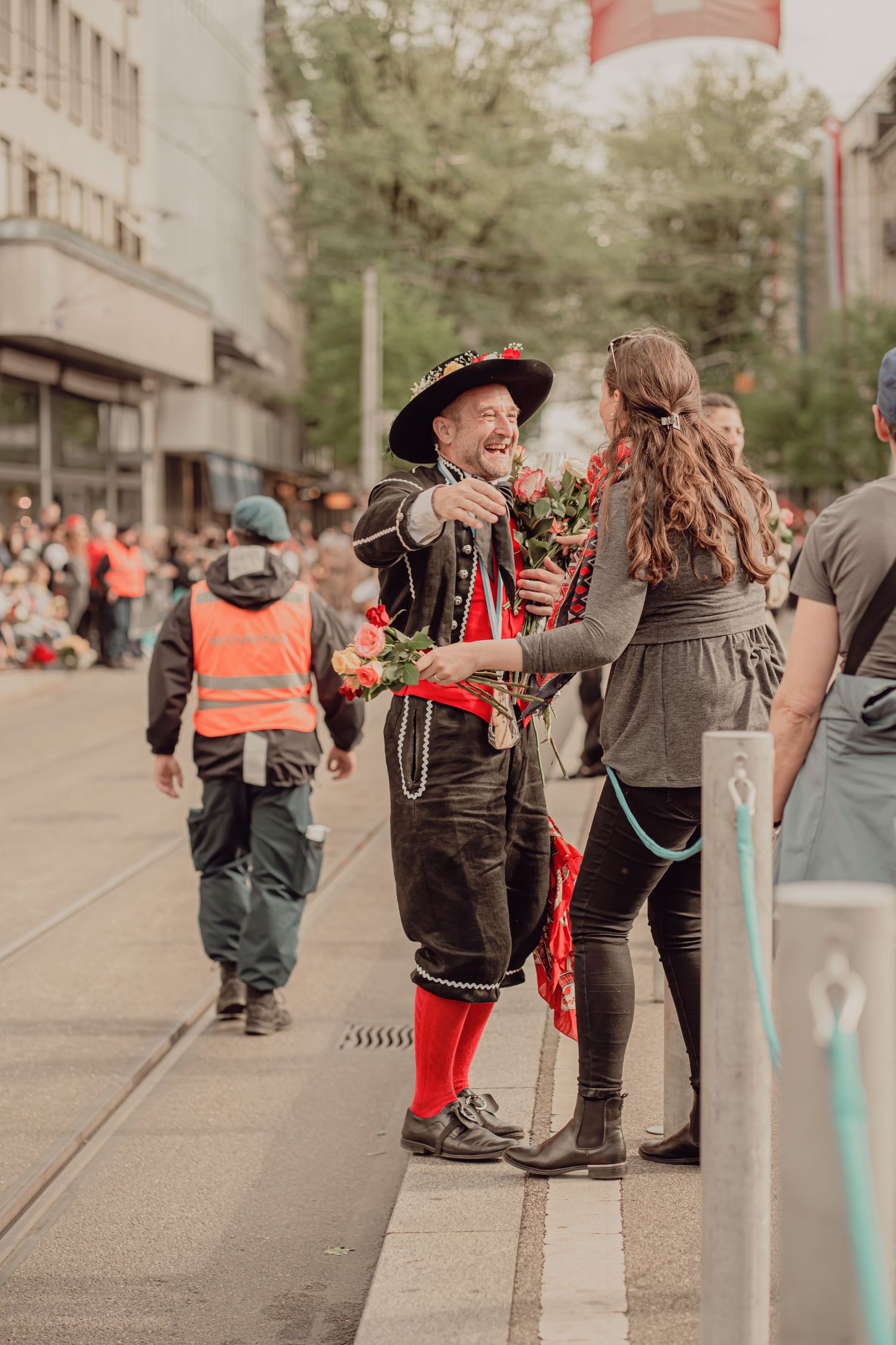 A cheerful man in traditional Swiss costume presents roses to a woman during a festive street parade.