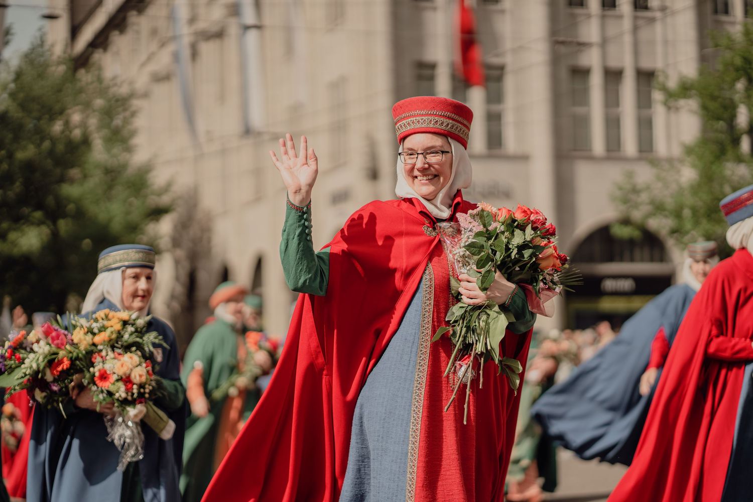 Smiling woman in red ceremonial cape waves to crowd while holding flowers in a street parade.