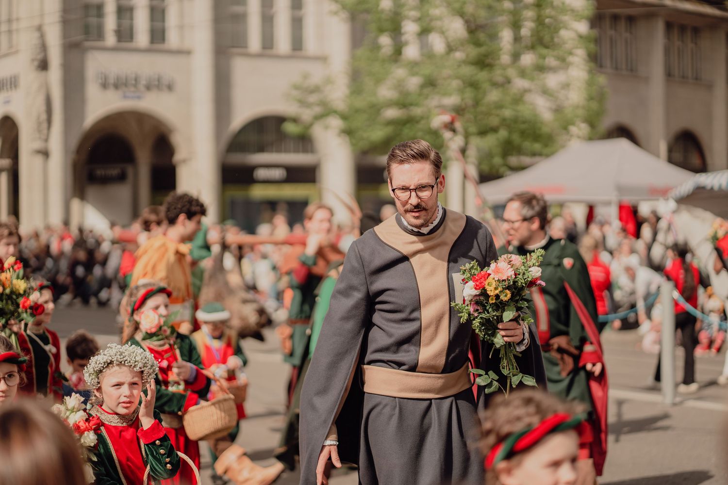 A man in medieval costume carries flowers while walking with children in red outfits during a Zürich parade.